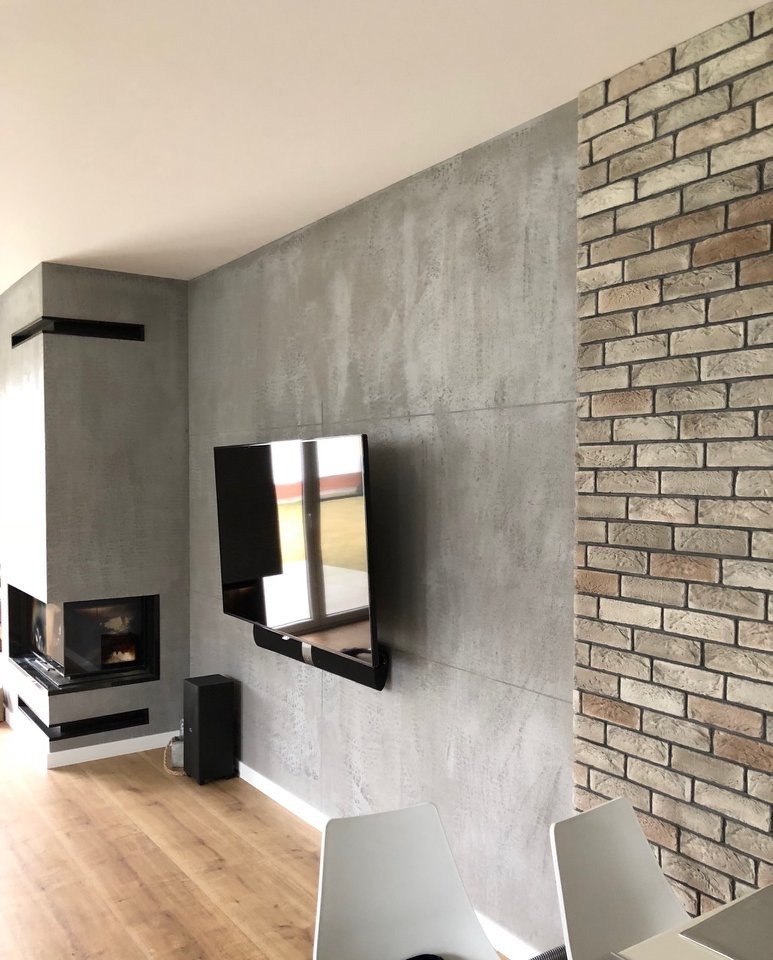 Wnętrze salonu z kominkiem, ścianą pokrytą dekoracyjną szarą fakturą przypominającą beton i fragmentem ściany wyłożonej cegłą, podwieszony telewizor, drewniana podłoga i białe krzesła.