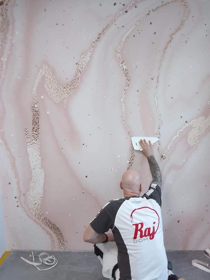 Pracownik w koszulce firmowej Raj Dom nakłada gładź imitującą marmur na ścianę za pomocą szpachelki, widoczne drobne niedoskonałości i resztki materiału na podłodze.