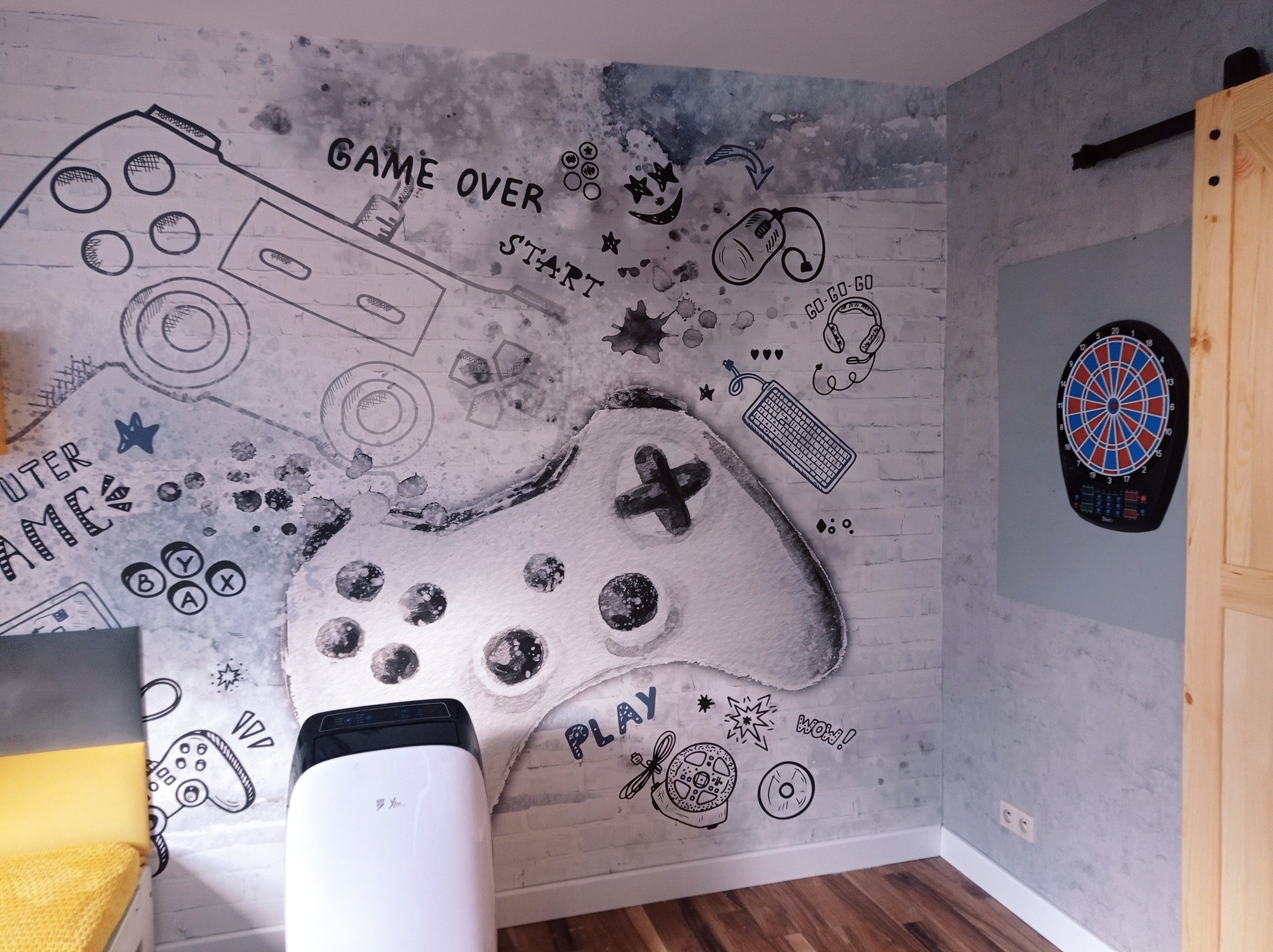 Pokój młodzieżowy z fototapetą w stylu gamingowym, na ścianie rysunki joysticków, słuchawek, myszki i klawiatury, widoczny również fragment tarczy do darta i przenośny klimatyzator.
