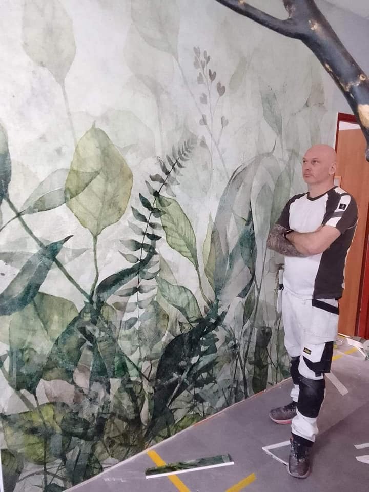 Mężczyzna z założonymi rękami stoi przed ścianą z motywem roślinnym w stylu akwareli, widoczne spodnie robocze i fragment podłogi z taśmą malarską.