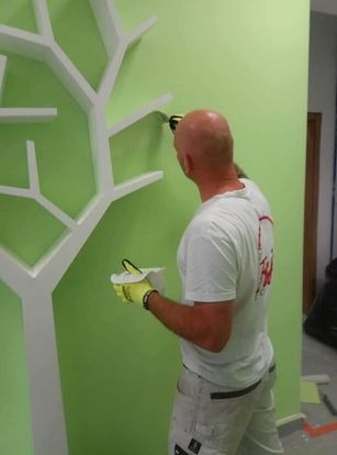 Malarz z Kórnika maluje jasnozieloną ścianę obok dekoracyjnego, białego drzewa ściennego, używając małego pędzla do precyzyjnego wykończenia.