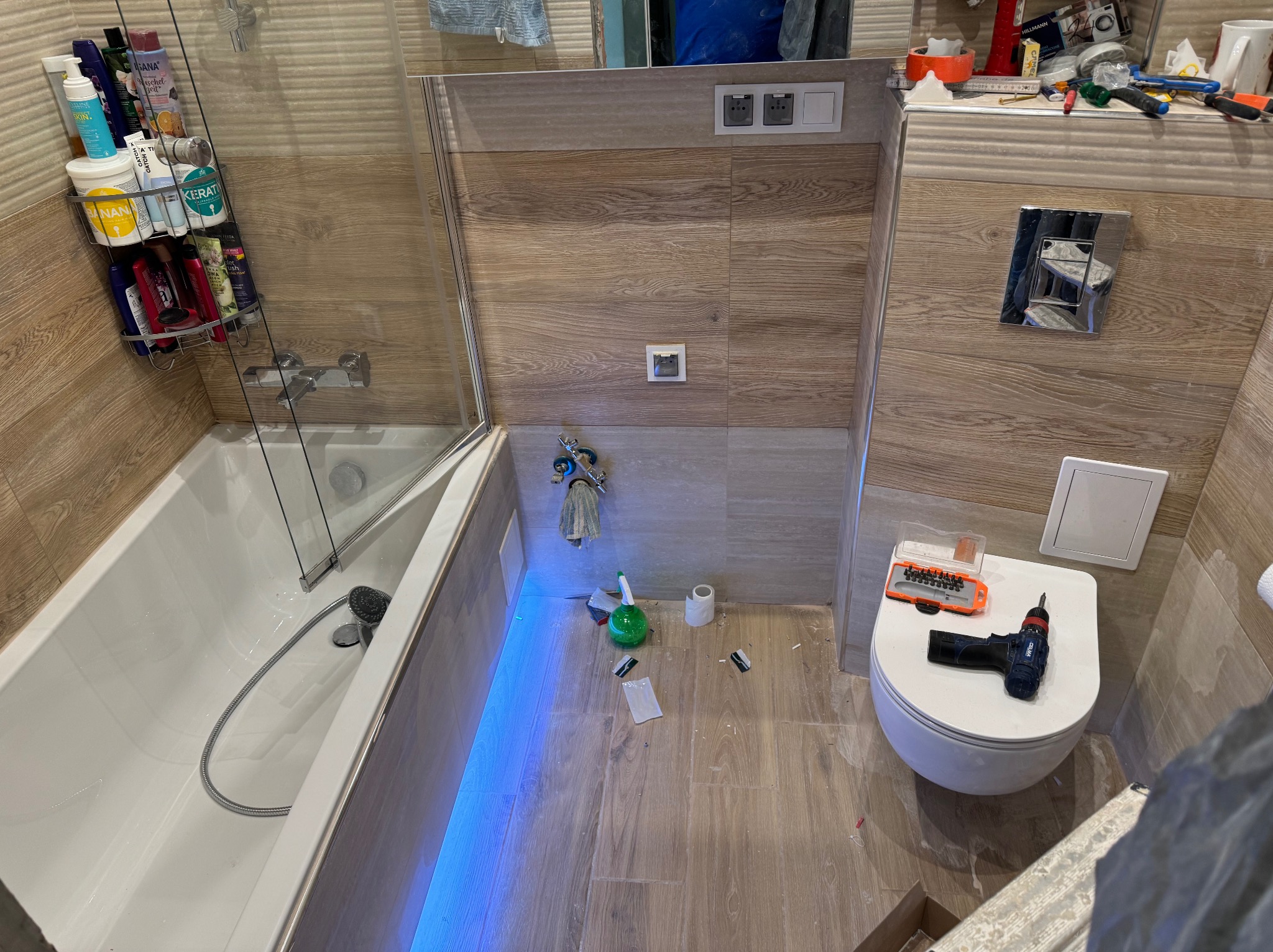 Niedokończona łazienka z wanną z parawanem, toaletą z narzędziami, płytkami imitującymi drewno i niebieskim podświetleniem LED wzdłuż wanny.