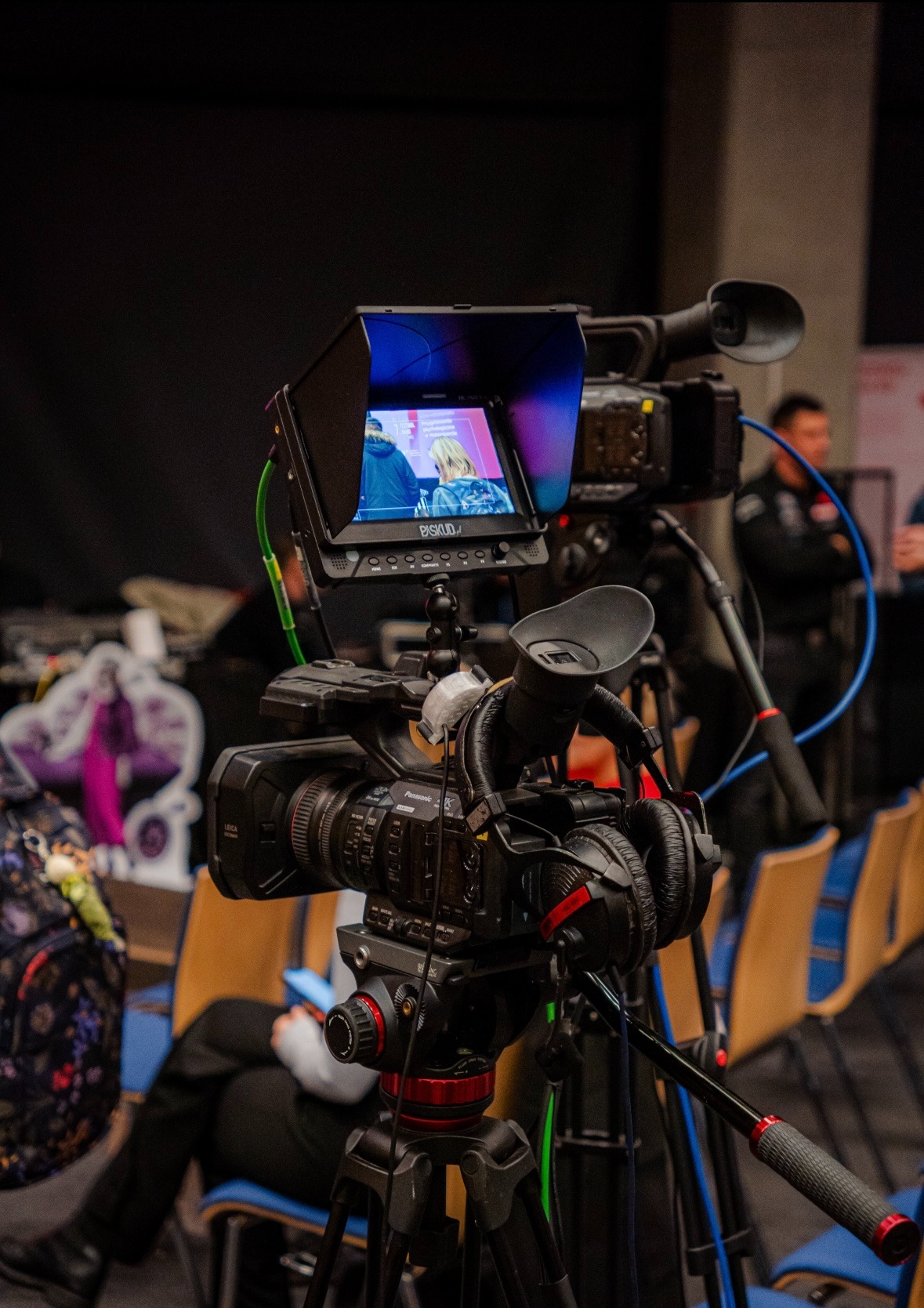 Profesjonalna kamera wideo Panasonic na statywie z założonymi słuchawkami, monitor podglądowy pokazuje nagrywany obraz, w tle inne kamery i osoby.