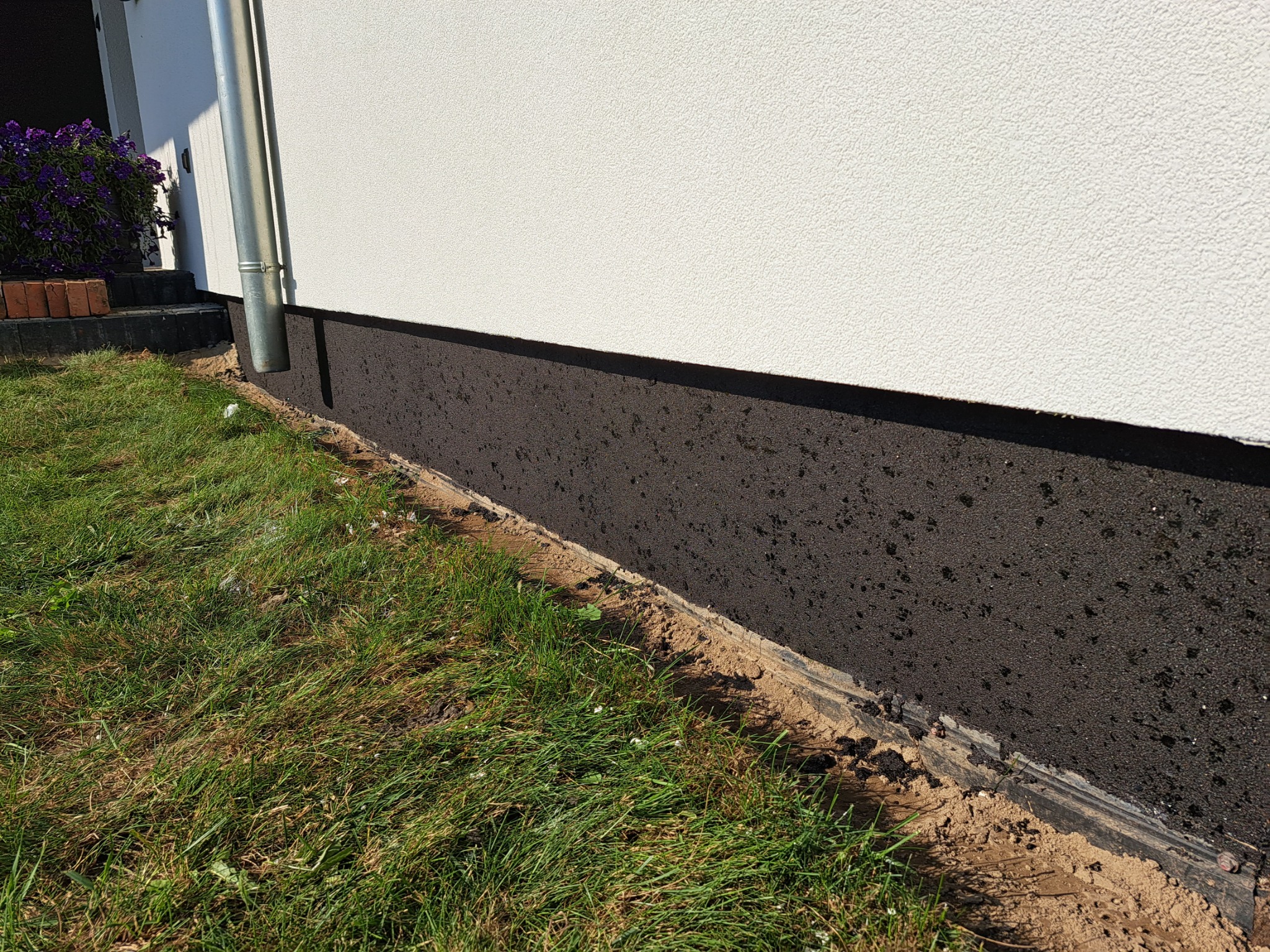 Dolna część elewacji budynku po zabezpieczeniu, z widoczną czarną, bitumiczną powłoką hydroizolacyjną, stykającą się z trawnikiem.