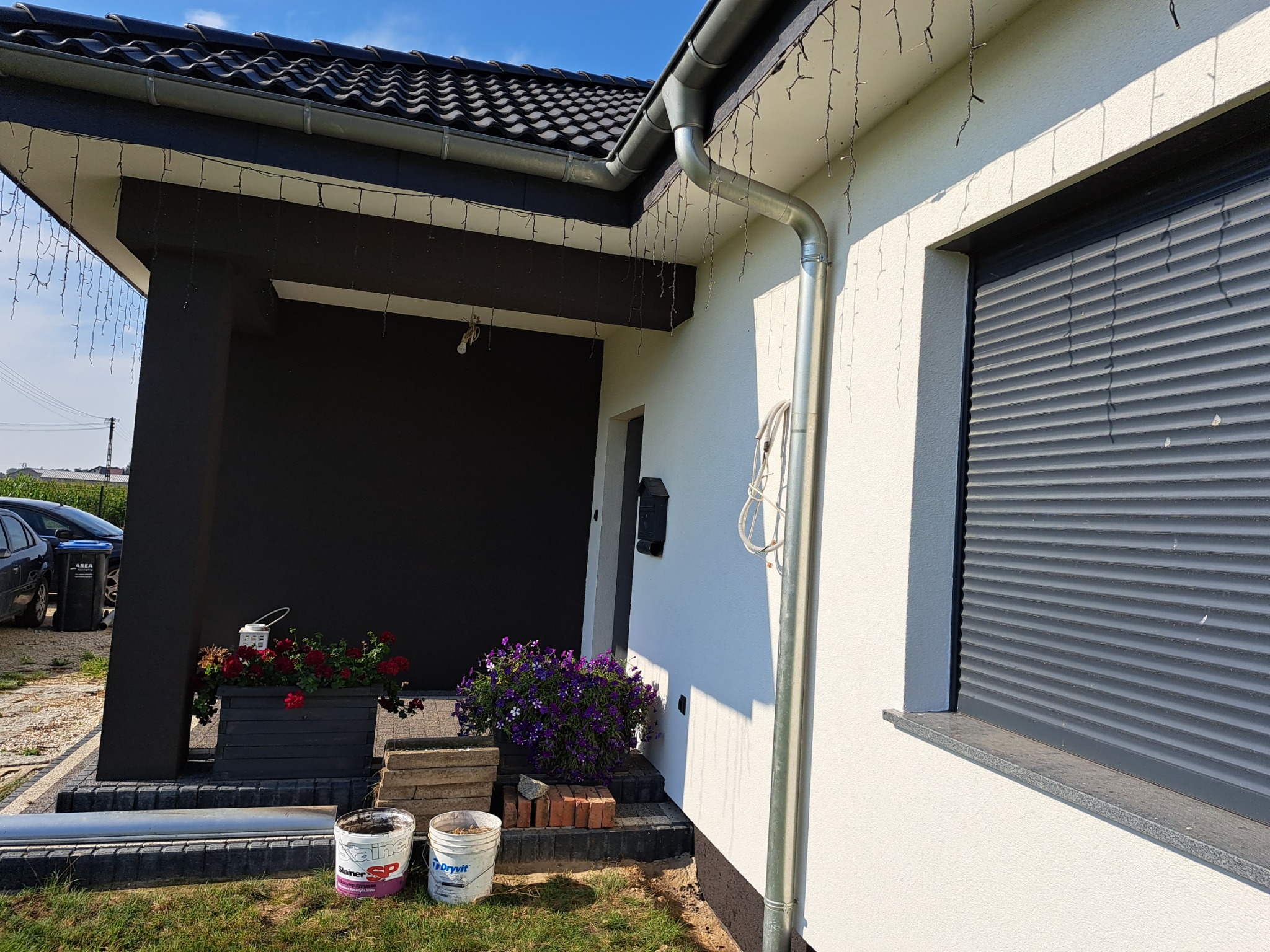 Elewacja domu jednorodzinnego po malowaniu, widoczne wiaderka z farbą, kwiaty w donicach i zwisające lampki dekoracyjne.