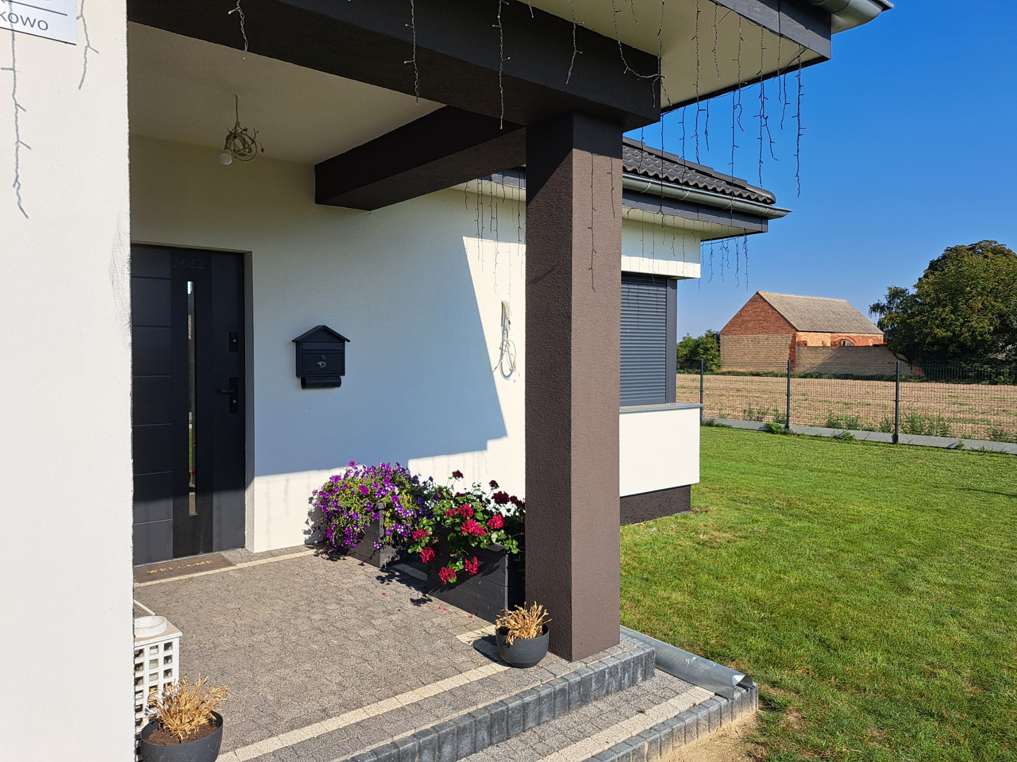 Wejście do domu jednorodzinnego z ciemnymi drzwiami i skrzynką na listy, ozdobione kwiatami w donicach, z widokiem na zadbany trawnik i budynek gospodarczy w tle.