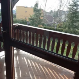 Ciemnobrązowa balustrada balkonu z widokiem na zieleń, jasne kafelki podłogowe z cieniami, drewniana rama okienna.