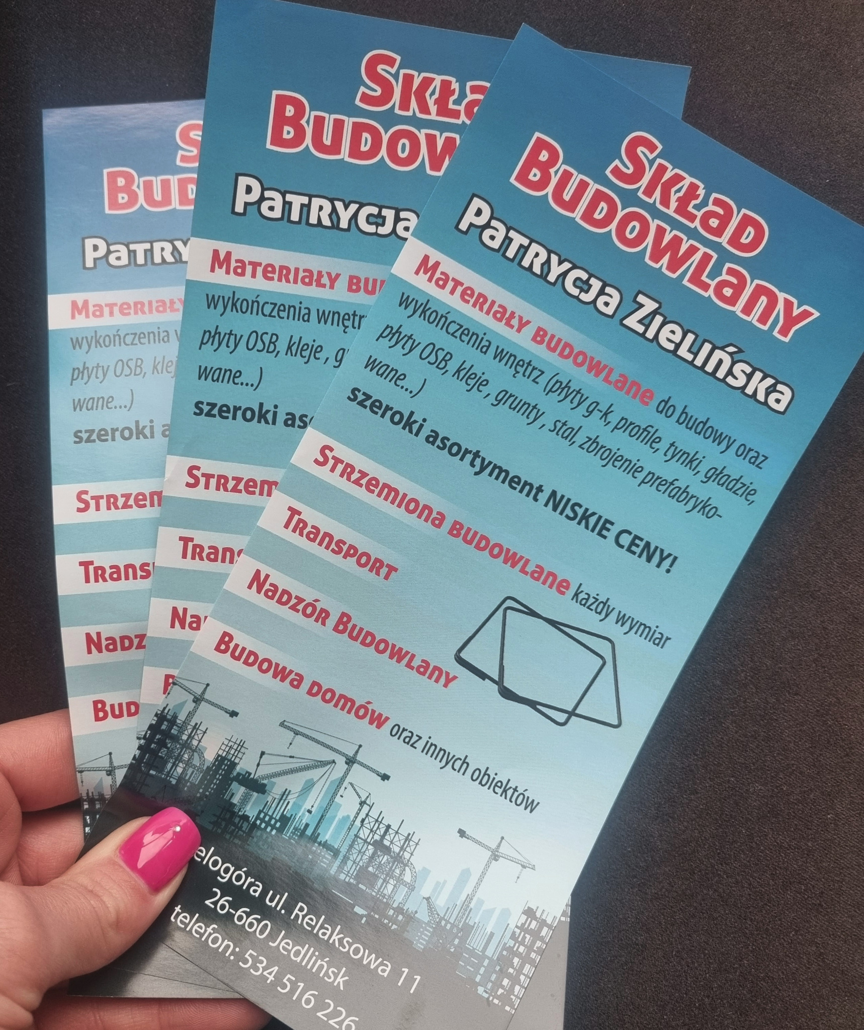 Dłoń z różowym lakierem na paznokciach trzyma trzy ulotki reklamowe firmy budowlanej z ofertą materiałów i usług, w tym transportu i nadzoru budowlanego, na ciemnym tle.