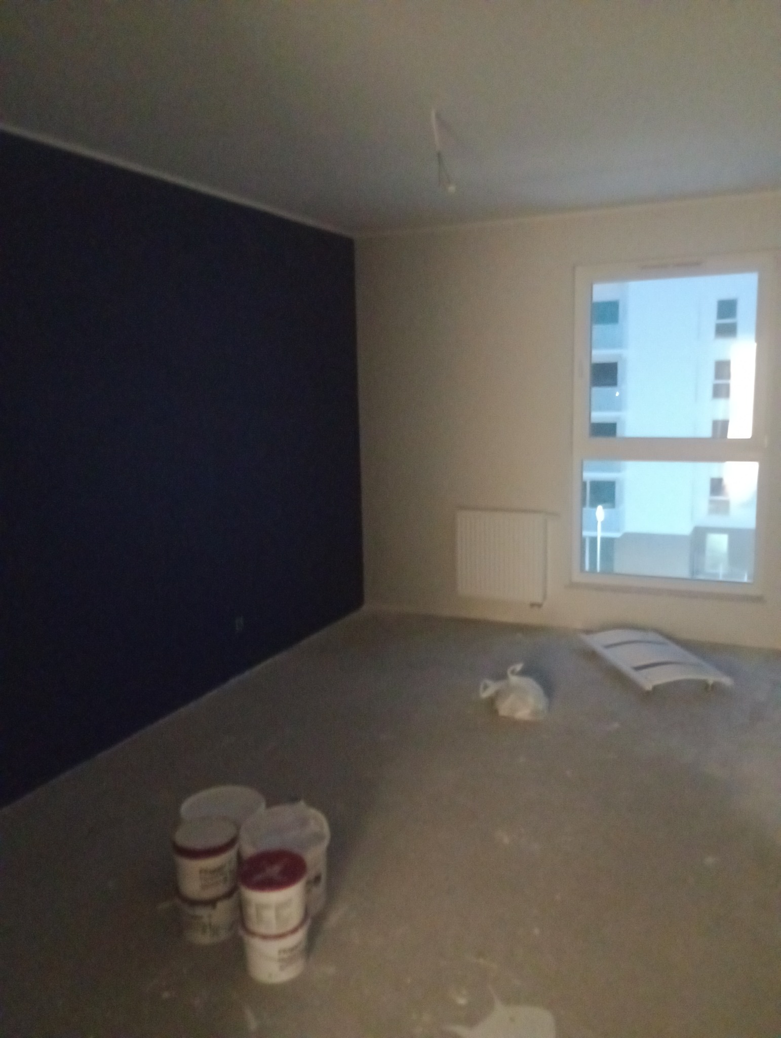 Pokój po częściowym pomalowaniu ścian na ciemny kolor, widoczne puszki z farbą, grzejnik i okno z widokiem na blok.