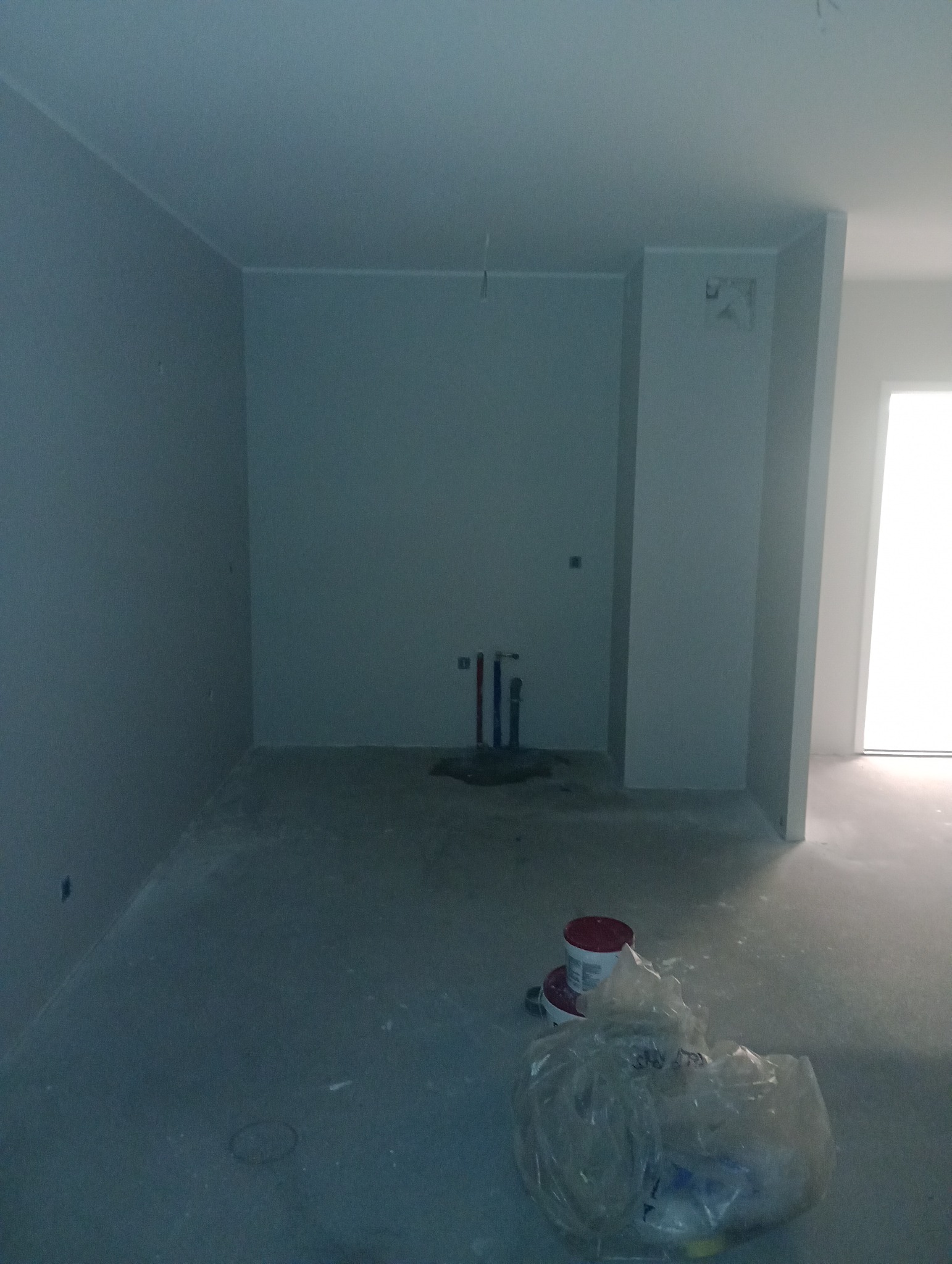 Wnętrze w trakcie prac wykończeniowych: świeżo pomalowane szare ściany, widoczne rury instalacji wodnej, puszki z farbą i worek z narzędziami na podłodze.