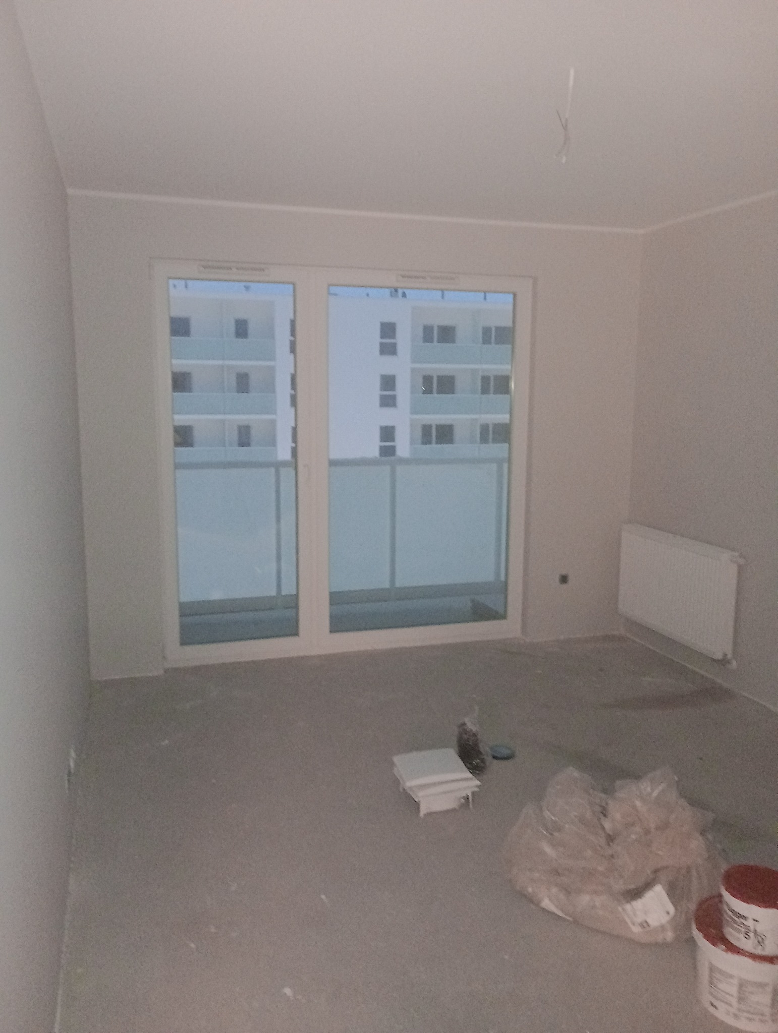 Pusty, świeżo otynkowany pokój w nowym budownictwie z widokiem na sąsiedni budynek przez balkonowe okno, widoczne wiaderka z farbą i akcesoria malarskie na podłodze.