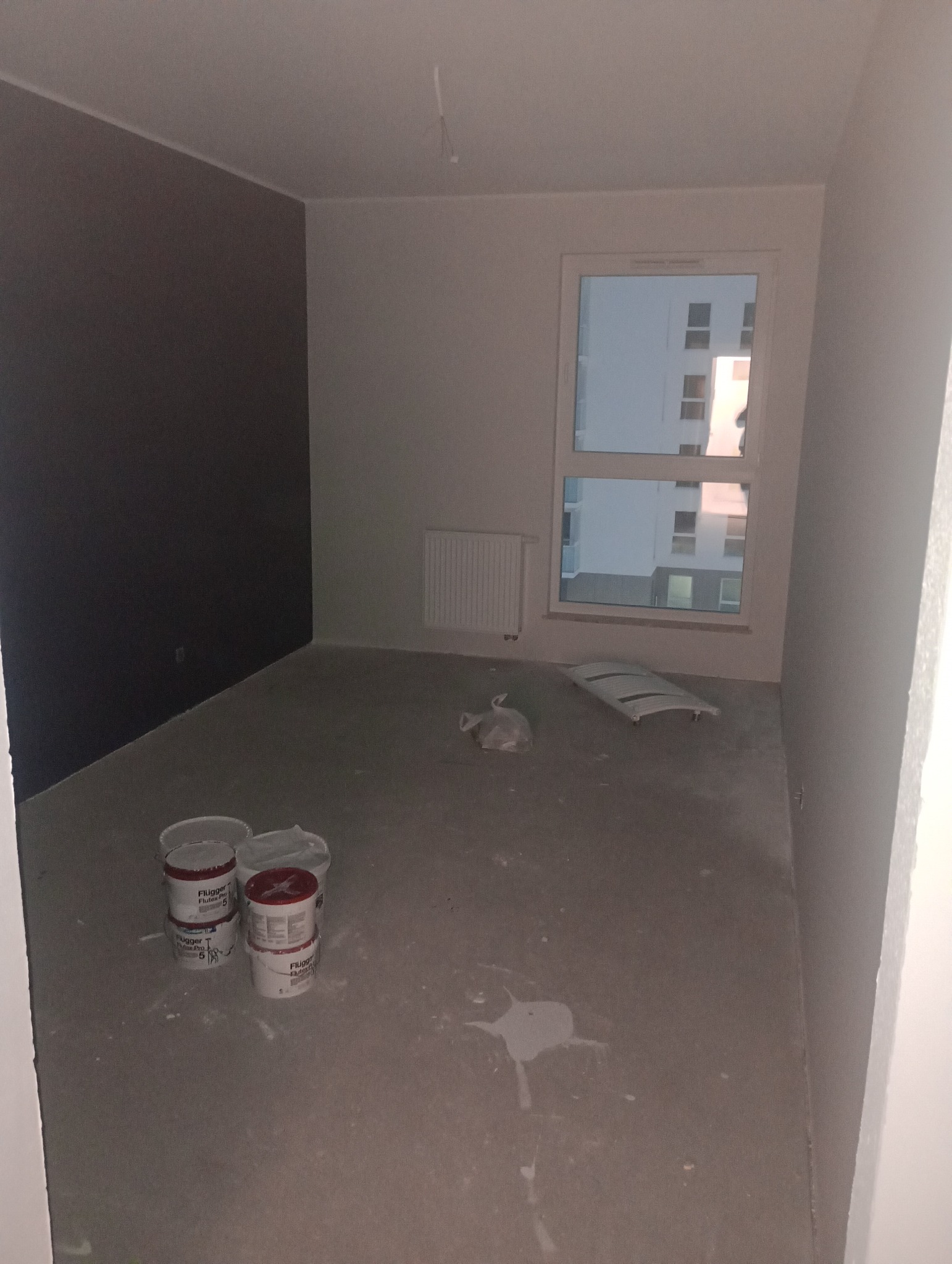 Pokój po malowaniu z widoczną ścianą pomalowaną na ciemny kolor, reszta ścian biała, widoczne puszki po farbie Flugger i resztki folii malarskiej na podłodze.