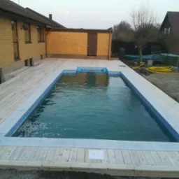 Prostokątny basen z mozaiką, otoczony jasnym drewnianym tarasem, przylegający do ż&oacute;łtego budynku z cegły, widok z g&oacute;ry.