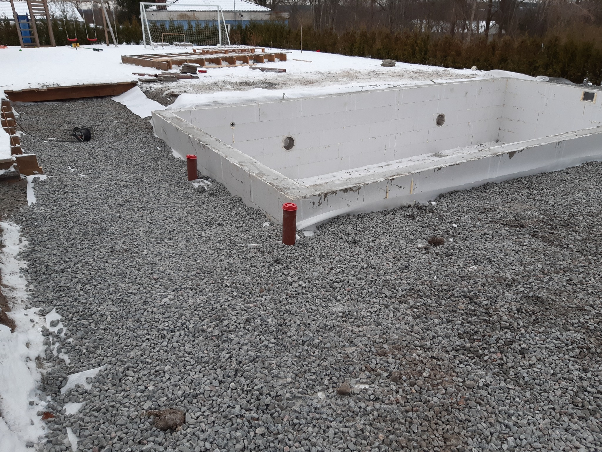 Wykop pod budowę basenu z widocznymi elementami konstrukcyjnymi z bloczków styropianowych i rurami odpływowymi, otoczony żwirem i częściowo pokryty śniegiem, w tle plac zabaw i boisko.