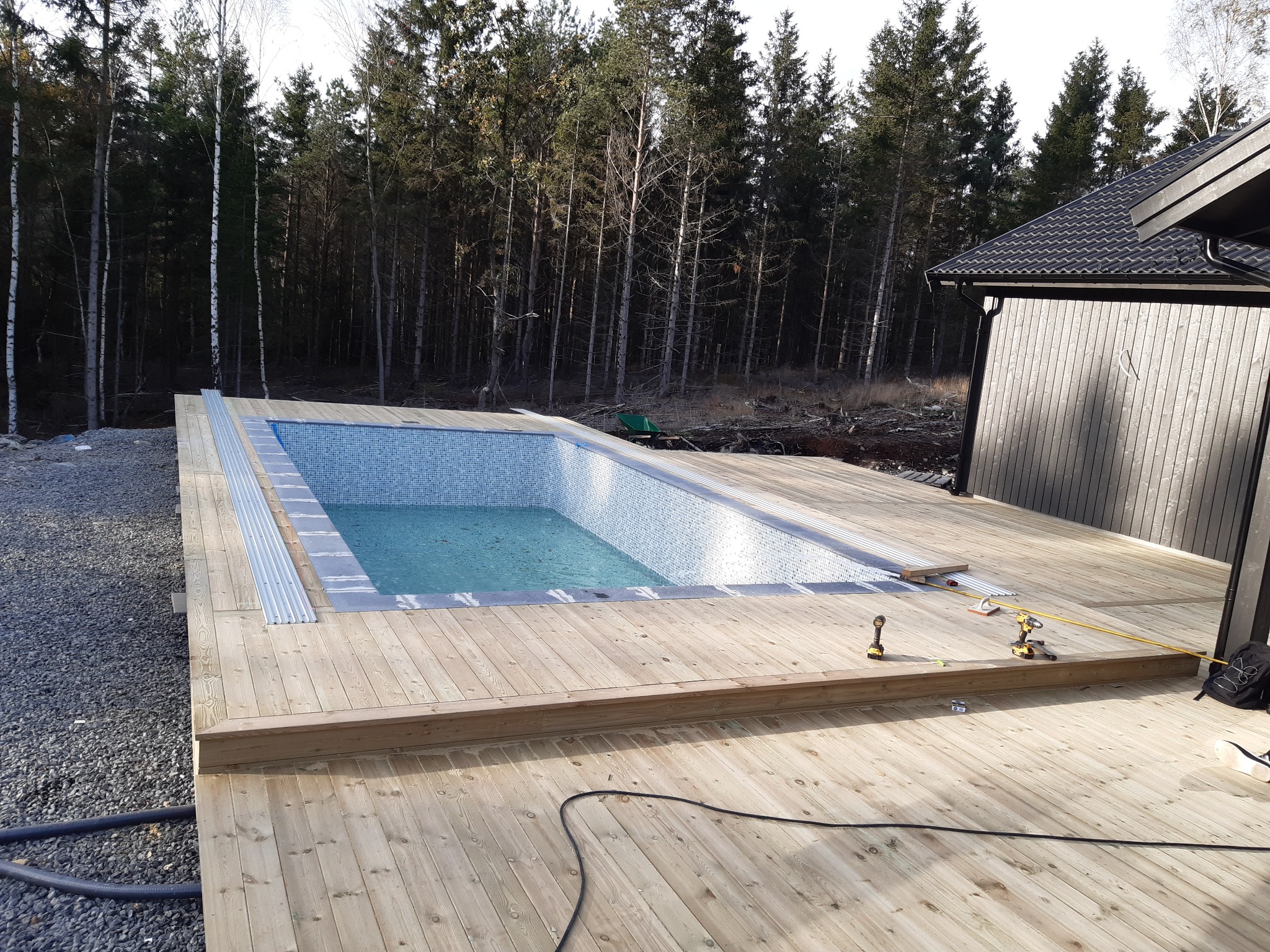 Nowoczesny basen z mozaiką wykończony drewnianym tarasem w trakcie budowy, z narzędziami i niezabezpieczonym brzegiem, na tle lasu i budynku z ciemną elewacją.