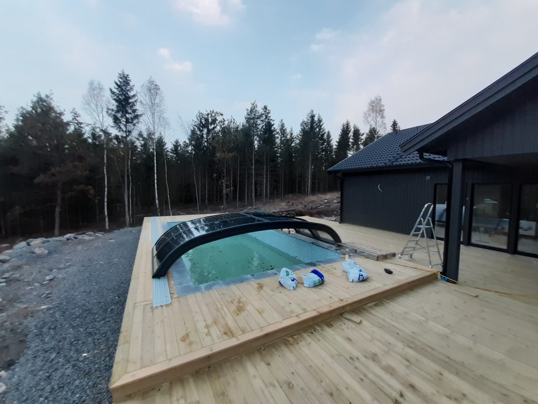 Zadaszony basen na drewnianym tarasie przylegającym do ciemnego domu, widok na las w tle.