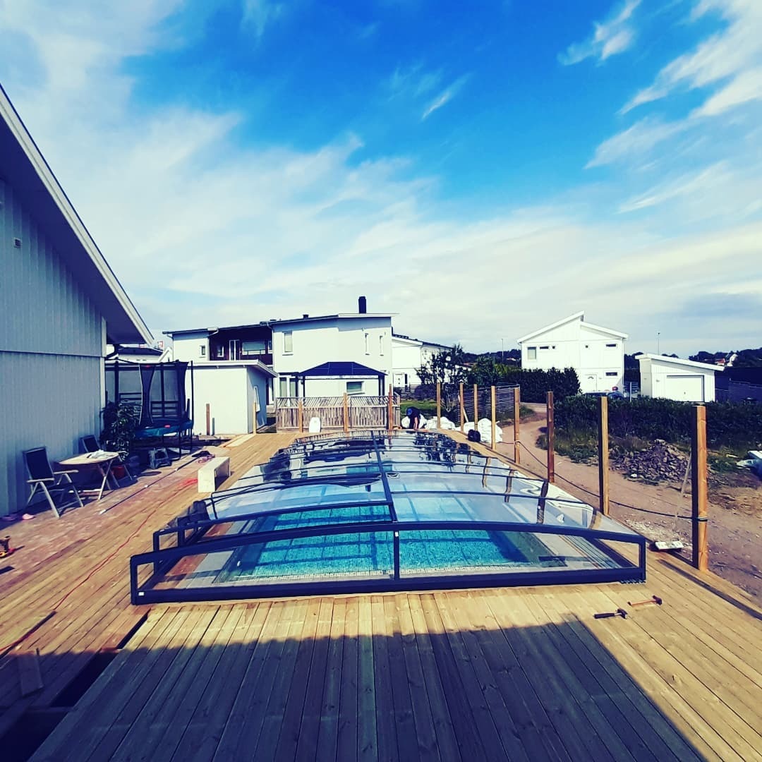 Zadaszony basen z drewnianym tarasem w trakcie budowy, widoczne narzędzia i elementy konstrukcyjne, w tle zabudowa jednorodzinna i błękitne niebo.