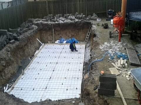 Przygotowanie podłoża pod budowę: wykop z ułożoną siatką zbrojeniową i izolacją, pracownik w odblaskowej kamizelce, betoniarka w tle.