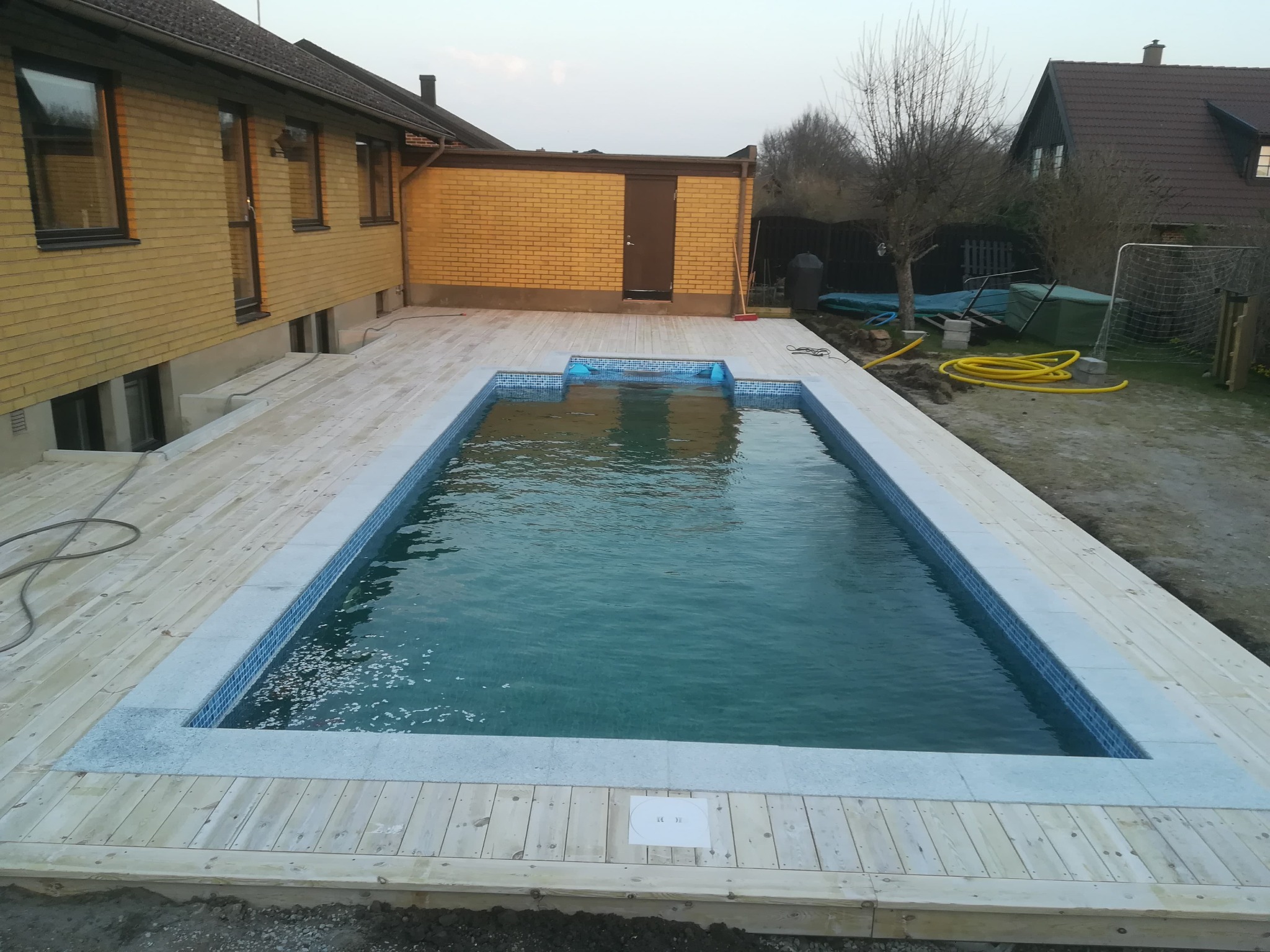 Prostokątny basen z mozaiką, otoczony jasnym drewnianym tarasem, przylegający do żółtego budynku z cegły, widok z góry.