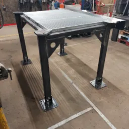 Stół spawalniczy z metalową kratą, czarne nogi z regulowanymi stopkami, widoczne ślady użytkowania w warsztacie.
