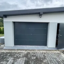 Zamknięta, segmentowa brama garażowa w kolorze antracytowym, z poziomymi przetłoczeniami i srebrnymi nitami, zamontowana w białym garażu z szarą podbitką, widok na kostkę brukową.