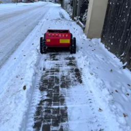 Czerwona maszyna do odśnieżania na chodniku z kostki brukowej, częściowo pokrytym śniegiem, z widocznymi śladami opon, w tle ulica i zabudowania w zimowej scenerii.