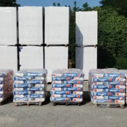 Na placu budowy składowane palety z zapakowanymi workami zaprawy murarskiej Solbet, białe płyty izolacyjne Solbet z czerwonym napisem 48/500 oraz cegły ceramiczne Cerpol w pomarańczowych opakowaniach.