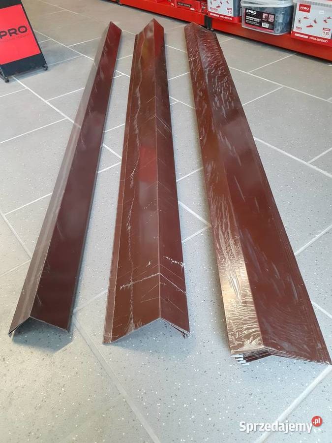 Trzy brązowe, metalowe profile kątowe, prawdopodobnie okapowe, ułożone na podłodze z płytek, częściowo owinięte folią ochronną.