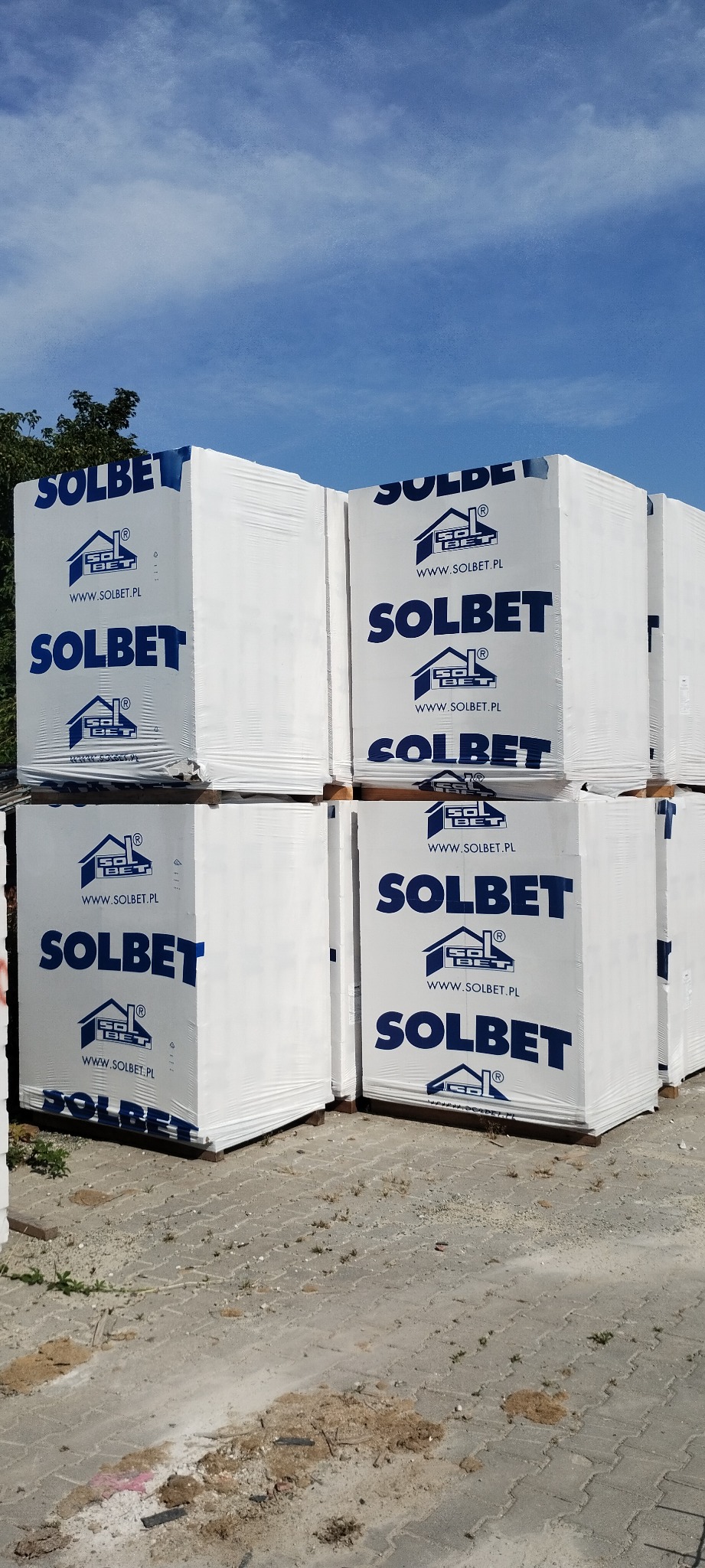 Palety z białymi paczkami materiału budowlanego SOLBET, zabezpieczone folią, na zewnątrz, w słoneczny dzień.