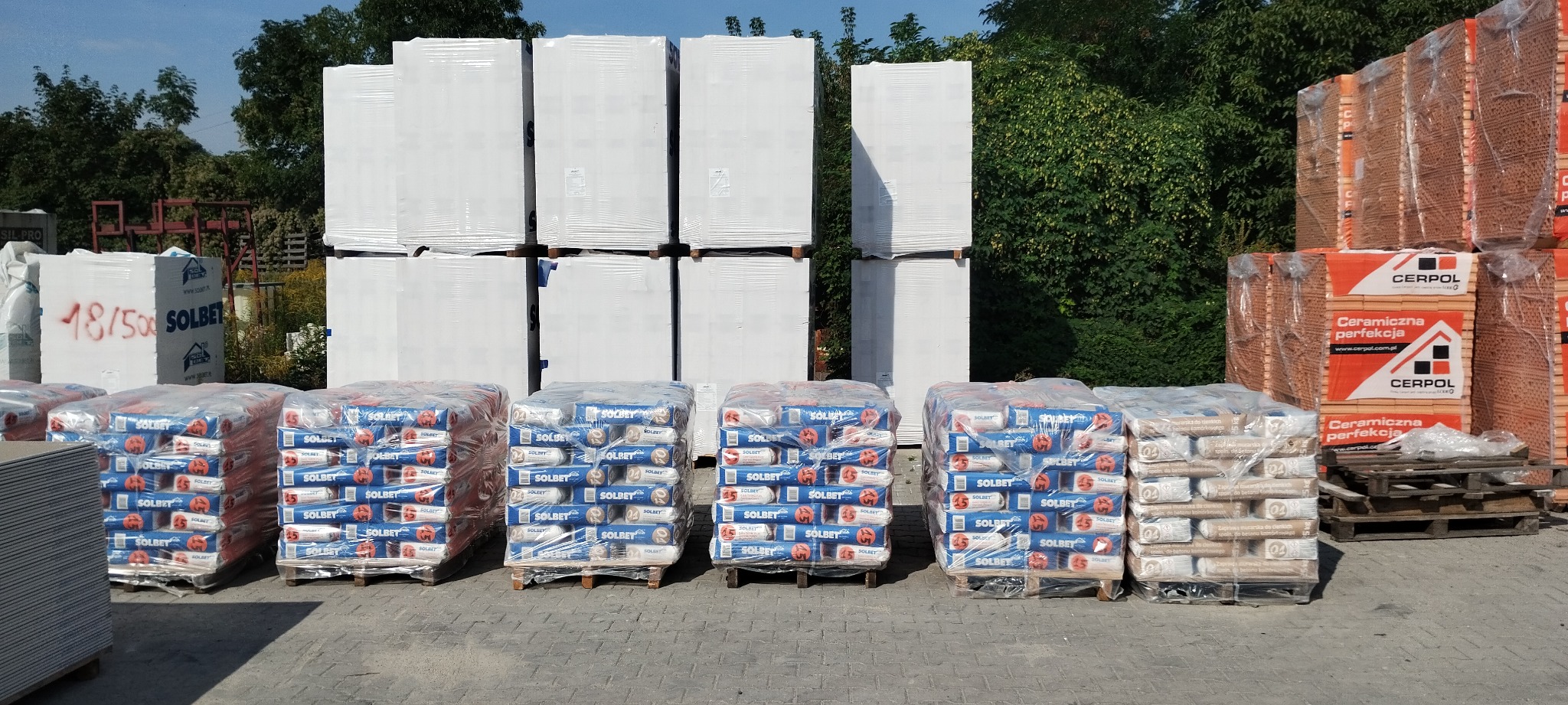 Na placu budowy składowane palety z zapakowanymi workami zaprawy murarskiej Solbet, białe płyty izolacyjne Solbet z czerwonym napisem 48/500 oraz cegły ceramiczne Cerpol w pomarańczowych opakowaniach.