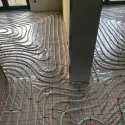 Białe rury ogrzewania podłogowego ułożone na folii z czerwoną siatką, przymocowane czarnymi uchwytami, widoczne w surowym stanie wnętrza z szarą kolumną i oknem w tle.