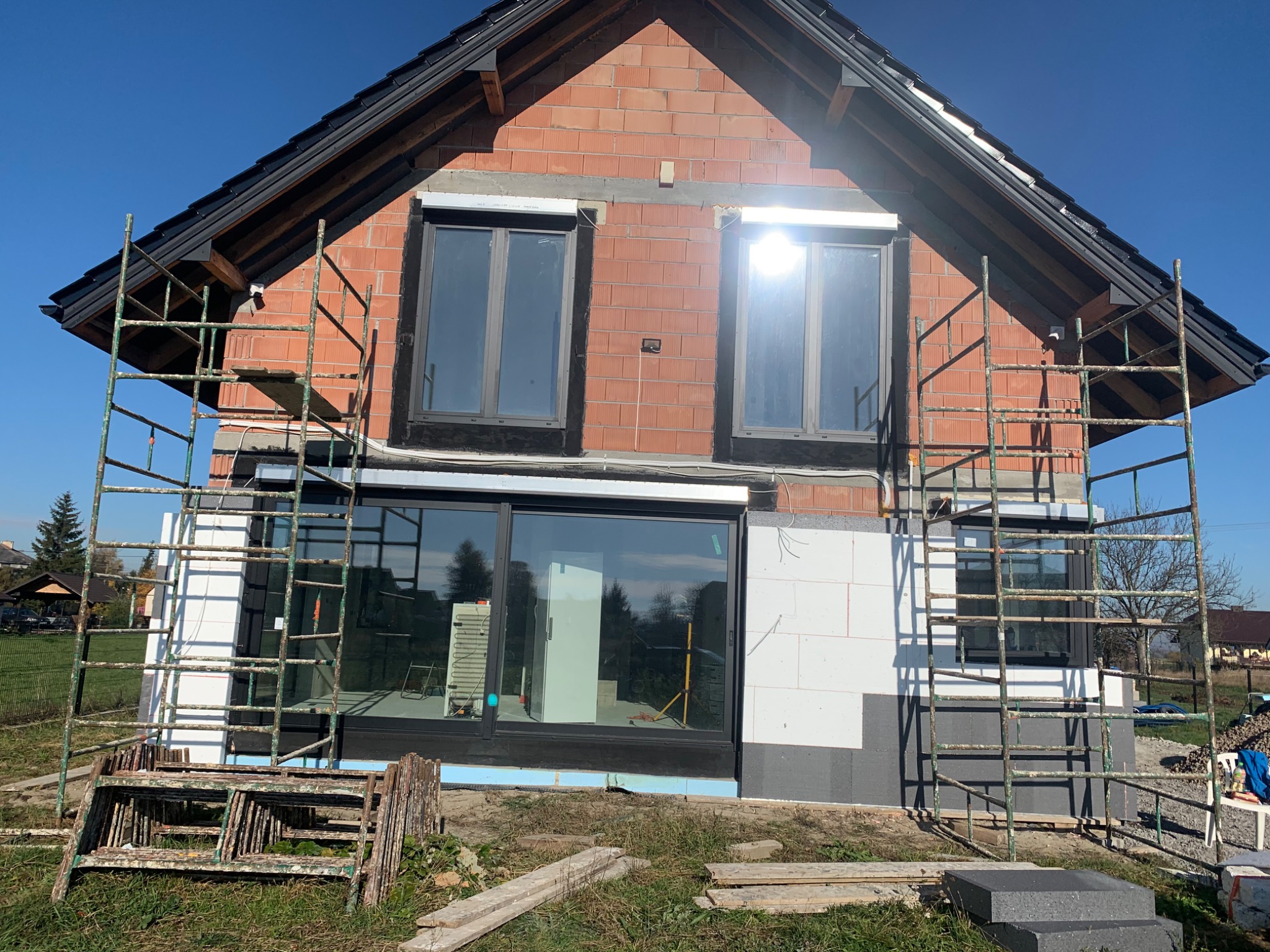 Surowy stan budynku w trakcie ocieplania, widoczne rusztowania, okna i fragmenty elewacji z cegły i styropianu.