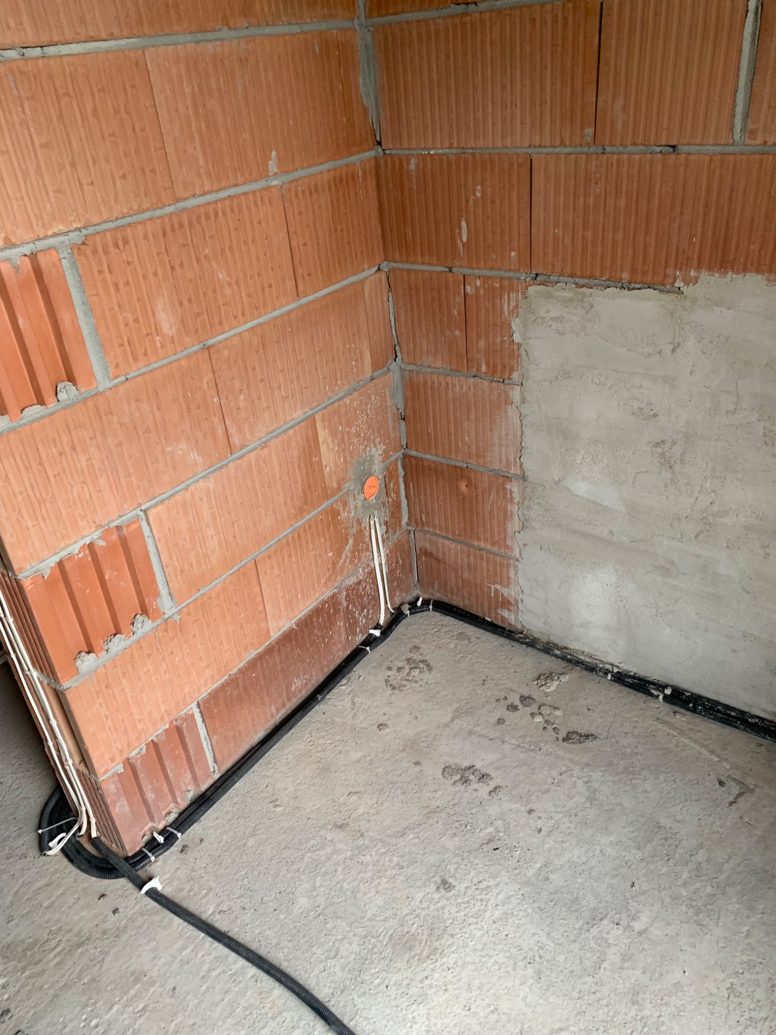 Fragment ściany z czerwonej cegły ceramicznej, częściowo otynkowanej, z widocznymi przewodami elektrycznymi w peszlach i rurkach, wychodzącymi ze ściany i biegnącymi wzdłuż podłogi z szarego betonu.