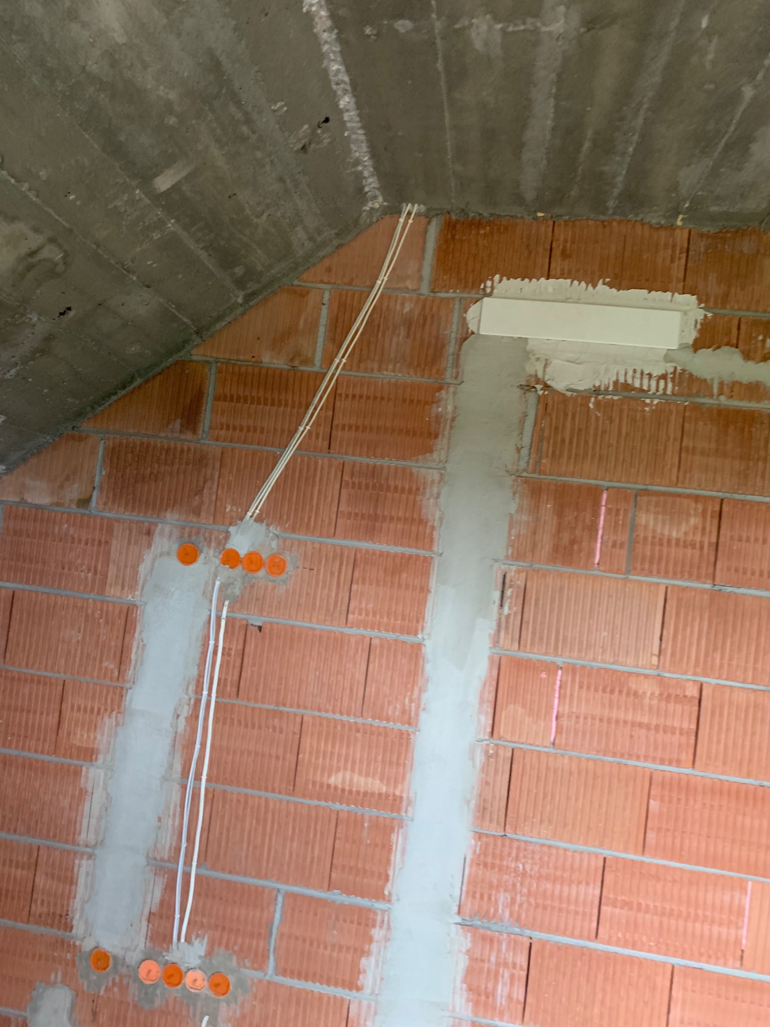 Surowa ściana z czerwonej cegły dziurawki, z bruzdami wypełnionymi szarym cementem, w których poprowadzone są białe kable elektryczne i pomarańczowe puszki instalacyjne, betonowy sufit pod skosem.