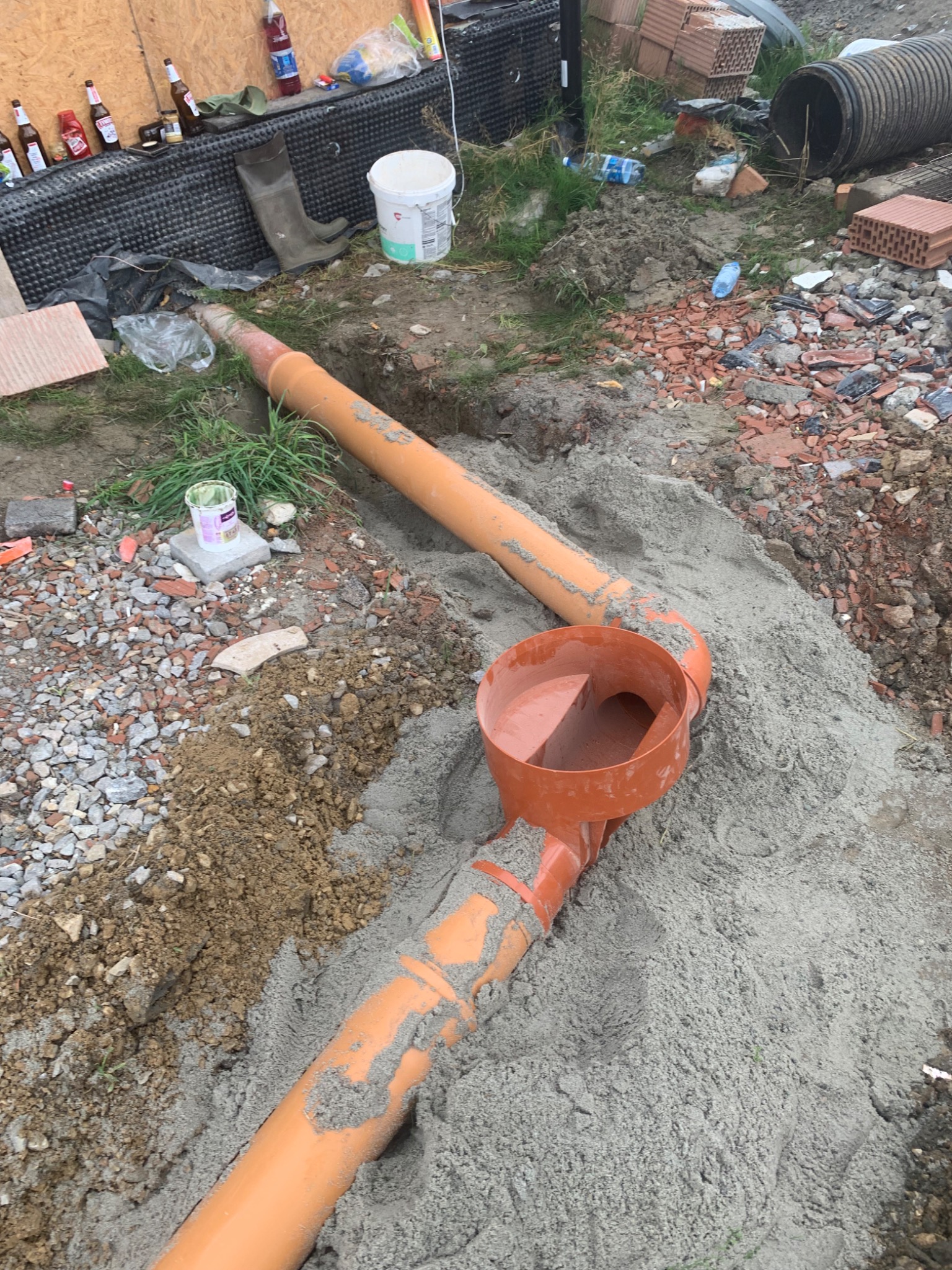 Pomarańczowe rury kanalizacyjne z kształtką inspekcyjną ułożone w wykopie, zasypywane piaskiem, widoczne gruz i materiały budowlane w tle.