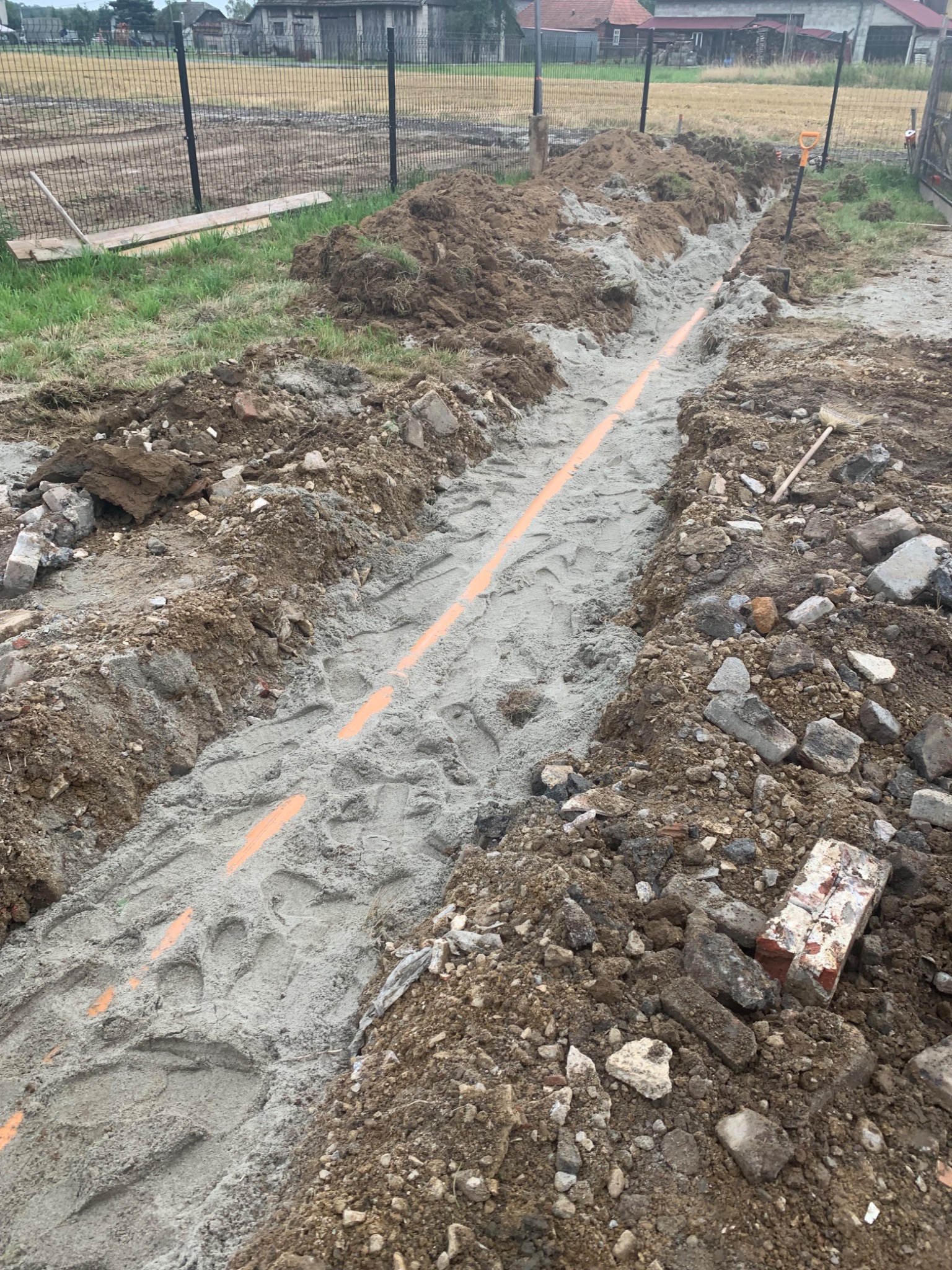 Wykop w ziemi z ułożonymi pomarańczowymi rurami, widoczne ślady stóp w piasku i ziemi, gruz i narzędzia na obrzeżach wykopu, w tle ogrodzenie i budynki.
