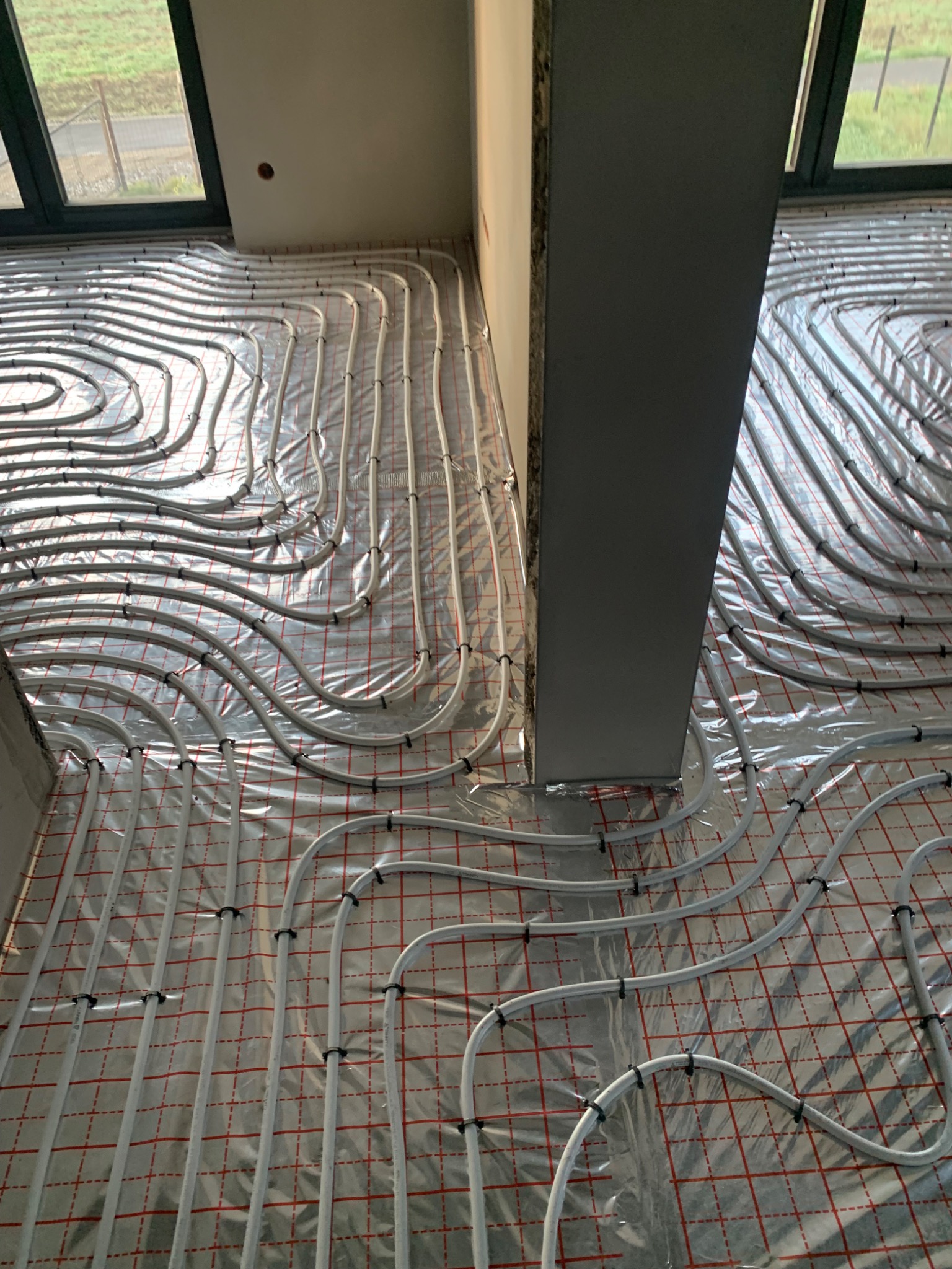 Białe rury ogrzewania podłogowego ułożone na folii z czerwoną siatką, przymocowane czarnymi uchwytami, widoczne w surowym stanie wnętrza z szarą kolumną i oknem w tle.