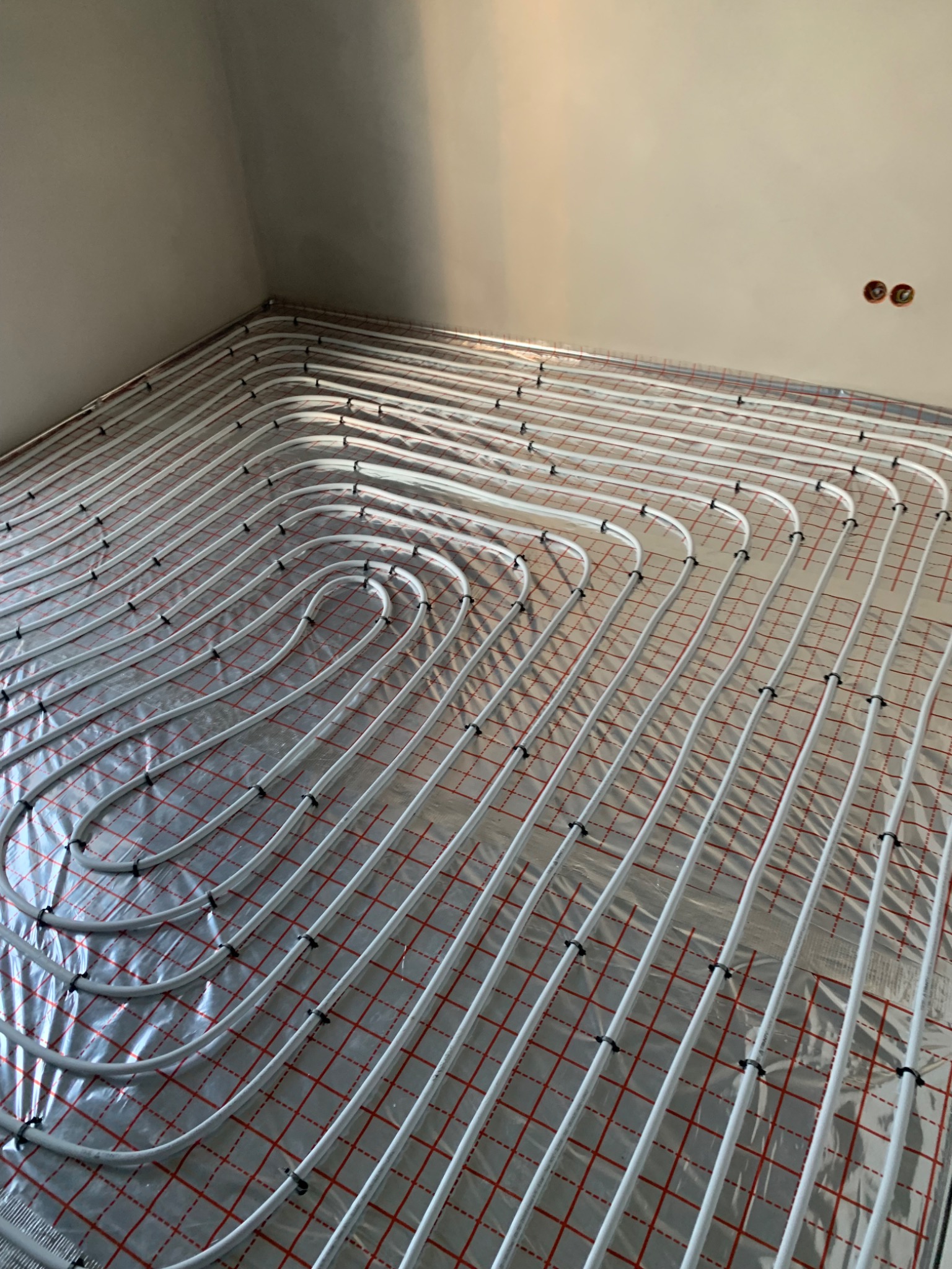 Białe rurki ogrzewania podłogowego ułożone na folii z czerwoną kratką w rogu jasnego pokoju, widoczne dwa otwory na ścianie.