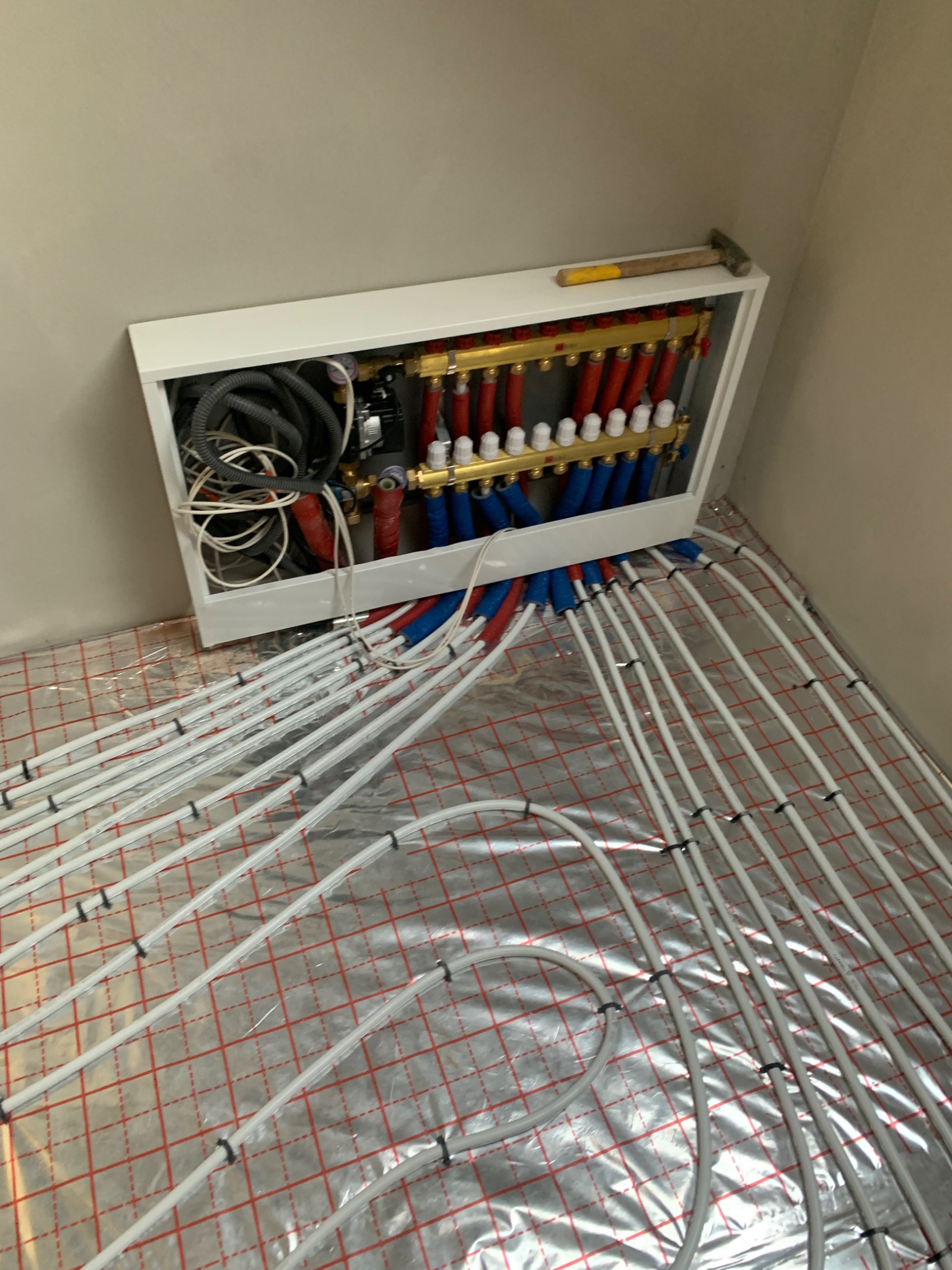 Rozdzielacz ogrzewania podłogowego z rurami ułożonymi na folii izolacyjnej z siatką, widoczny młotek na górnej krawędzi obudowy.