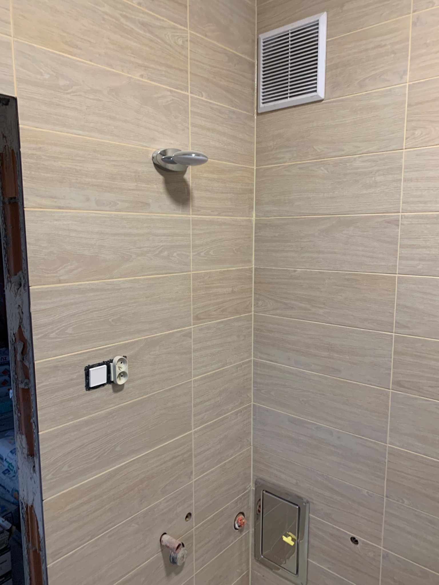 Ściana łazienki wyłożona beżowymi płytkami imitującymi drewno, z widocznym otworem wentylacyjnym, włącznikiem światła, uchwytem na ręcznik oraz otworem rewizyjnym w dolnej części. Widoczne ślady po...