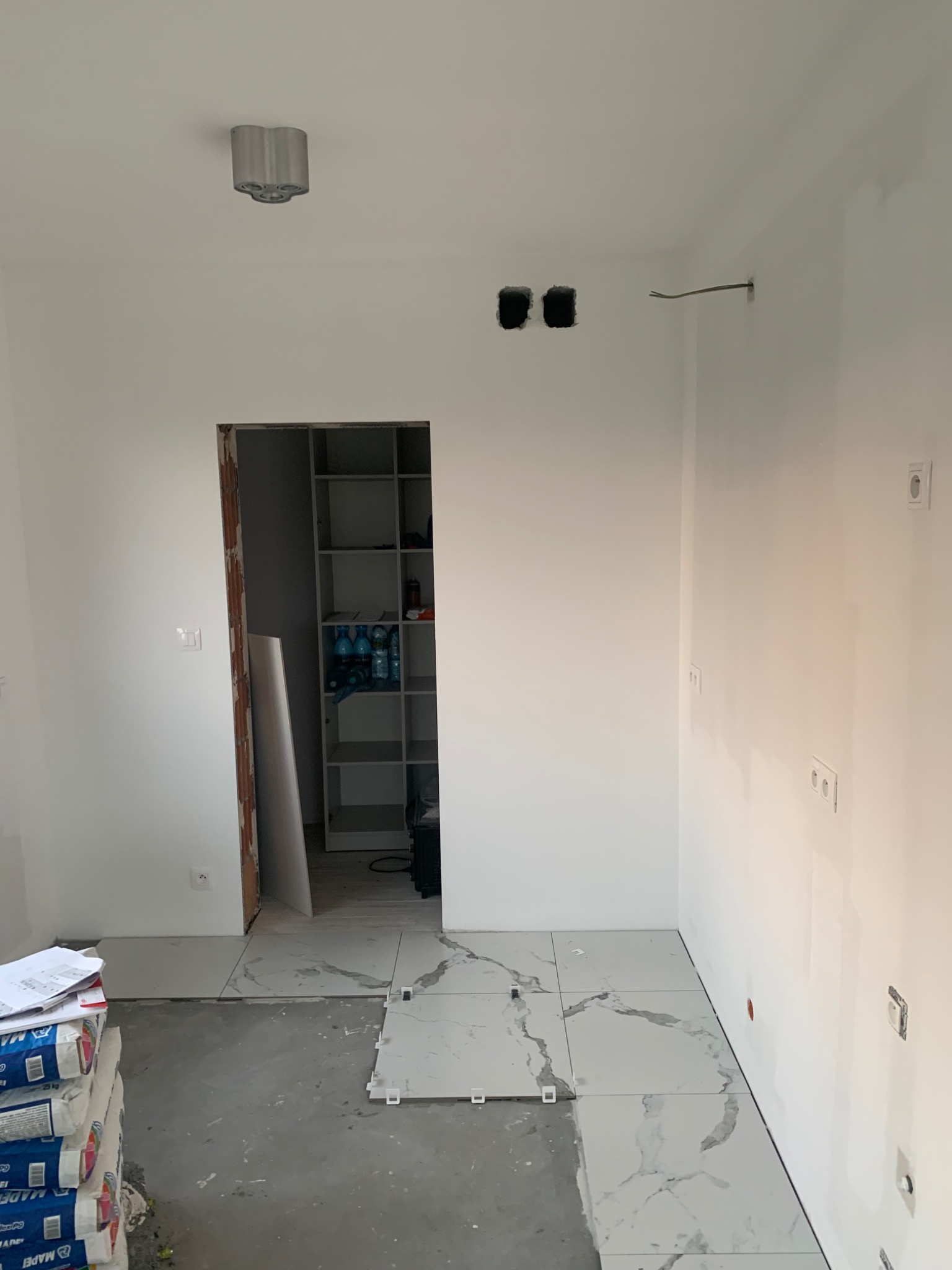 Wnętrze w trakcie układania płytek podłogowych z marmurowym wzorem; worki z klejem w rogu, widoczne puszki elektryczne i wyprowadzony przewód, surowa ościeżnica drzwiowa.
