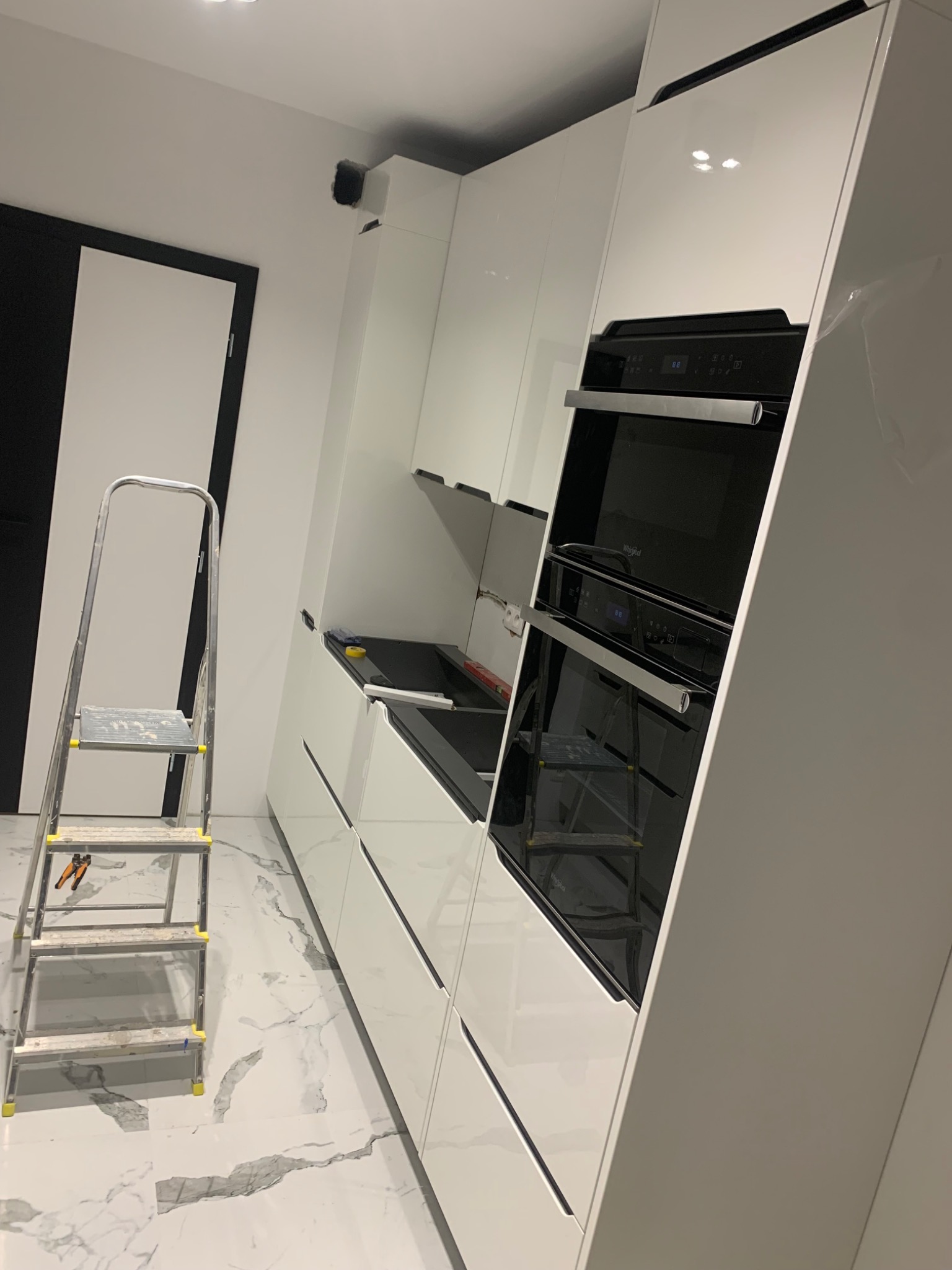 Nowoczesna kuchnia z białymi, błyszczącymi szafkami, marmurową podłogą, drabina malarska i wbudowany piekarnik Whirlpool podczas montażu.