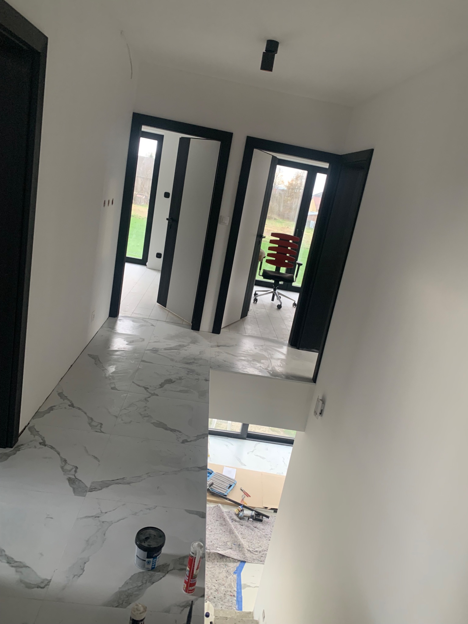 Wnętrze po wykończeniu: białe ściany, marmurowe płytki podłogowe, czarne ramy drzwi, widoczne puszki farb i narzędzia malarskie na podłodze, w tle biurowe krzesło.