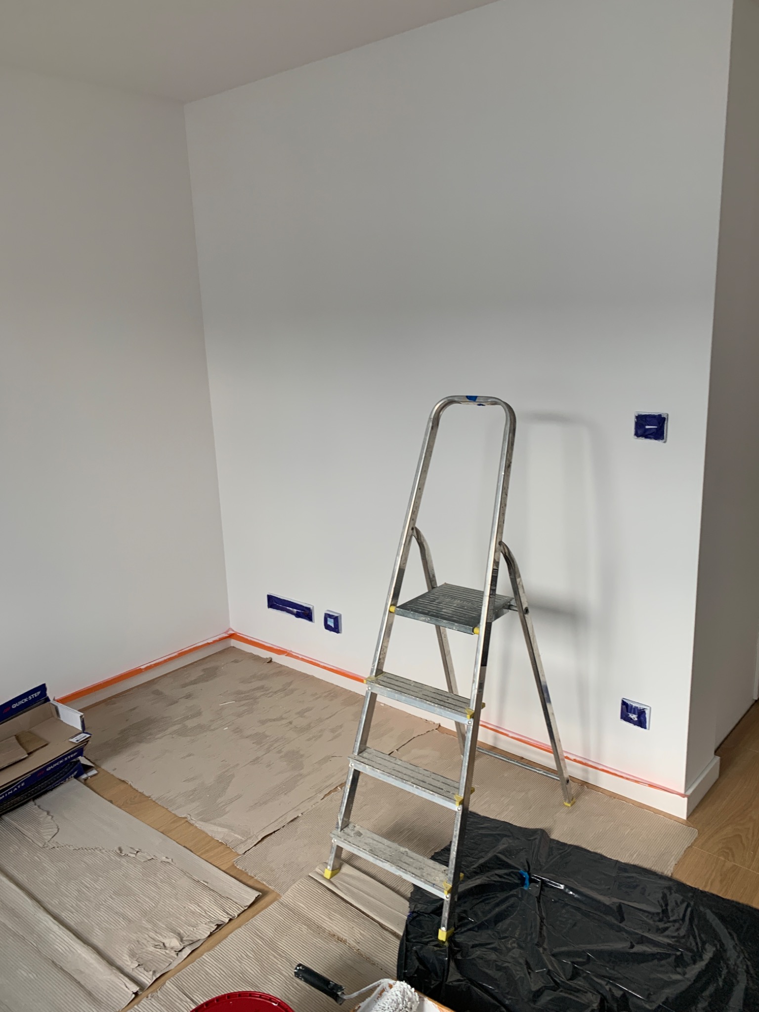 Pokój w trakcie malowania: białe ściany, zabezpieczone listwy przypodłogowe pomarańczową taśmą, zabezpieczona podłoga, aluminiowa drabina, zakryte gniazdka elektryczne niebieską taśmą, widoczny...