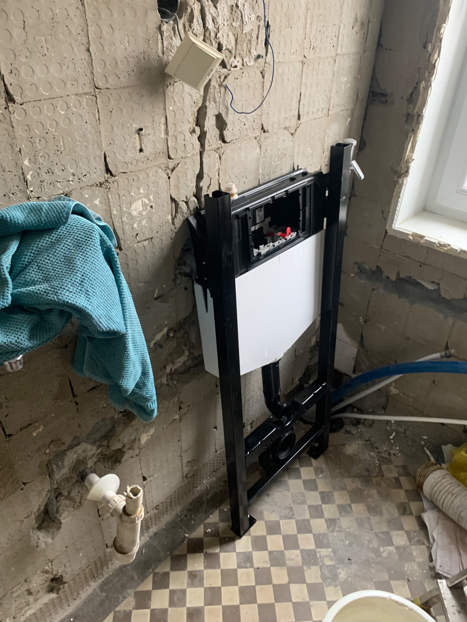 Stelaż podtynkowy WC zamontowany w łazience w trakcie remontu, widoczne instalacje wodne, stara, zniszczona ściana z resztkami płytek i wiszący turkusowy ręcznik.