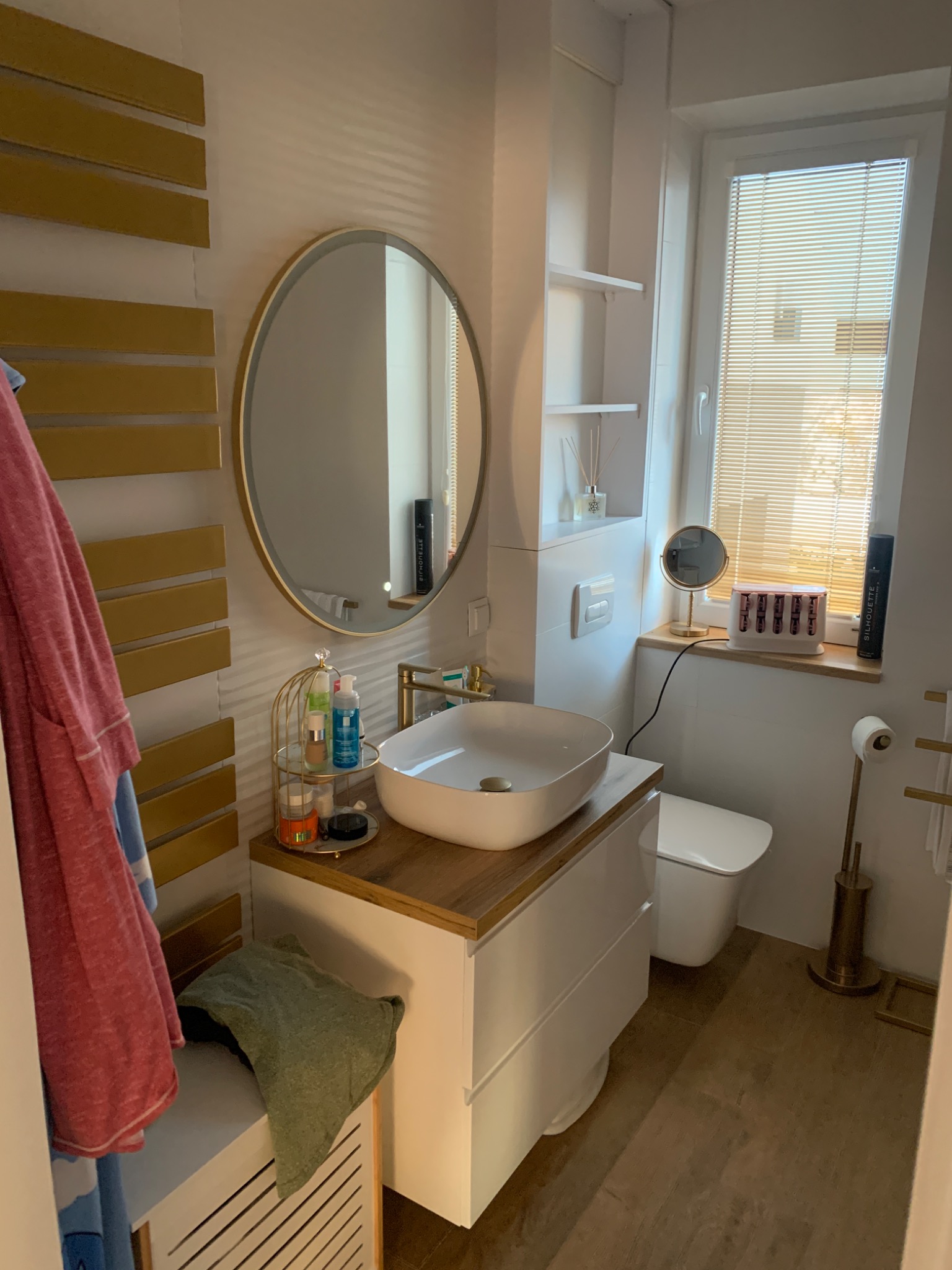 Nowoczesna łazienka z owalnym lustrem w złotej ramie, białą umywalką nablatową na szafce z drewnianym blatem, grzejnikiem drabinkowym w kolorze złotym oraz białymi płytkami i drewnopodobną podłogą.