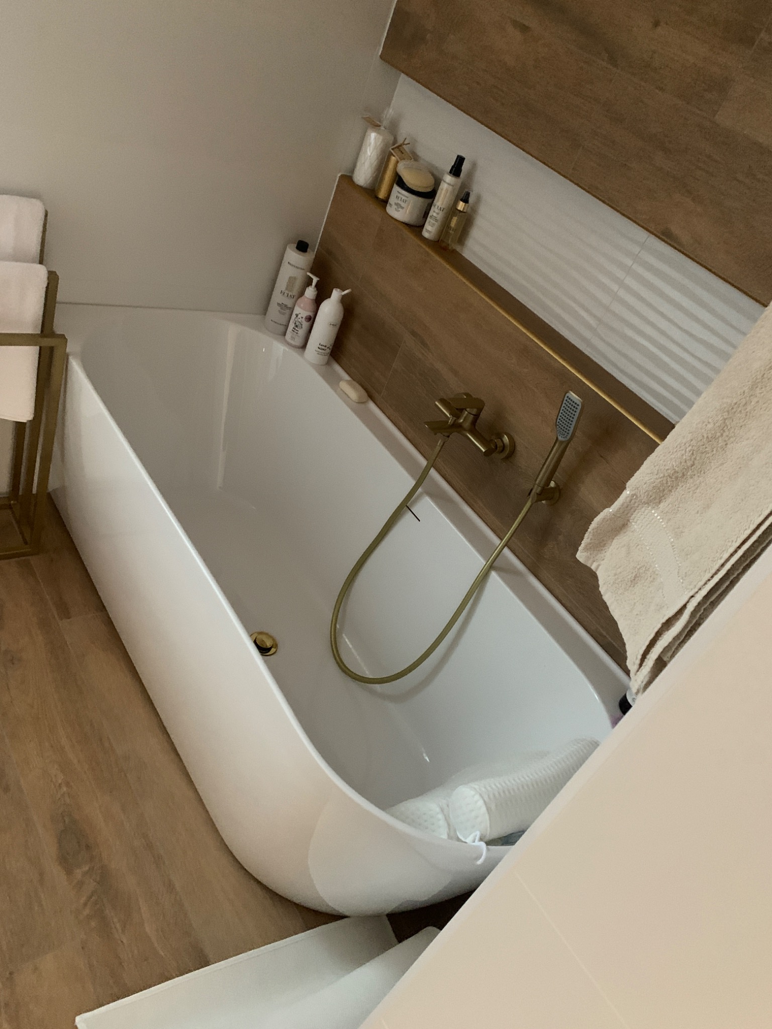 Nowoczesna, biała wanna wolnostojąca z złotą armaturą, drewnopodobna ściana w tle, minimalistyczne akcesoria łazienkowe na półce, ręcznik obok wanny.