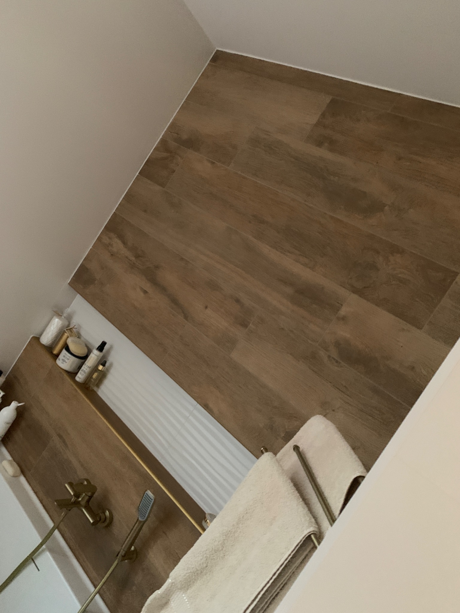 Ukośne ujęcie wykończonej łazienki z drewnopodobną podłogą, złotą armaturą i półką z kosmetykami, widok z góry.