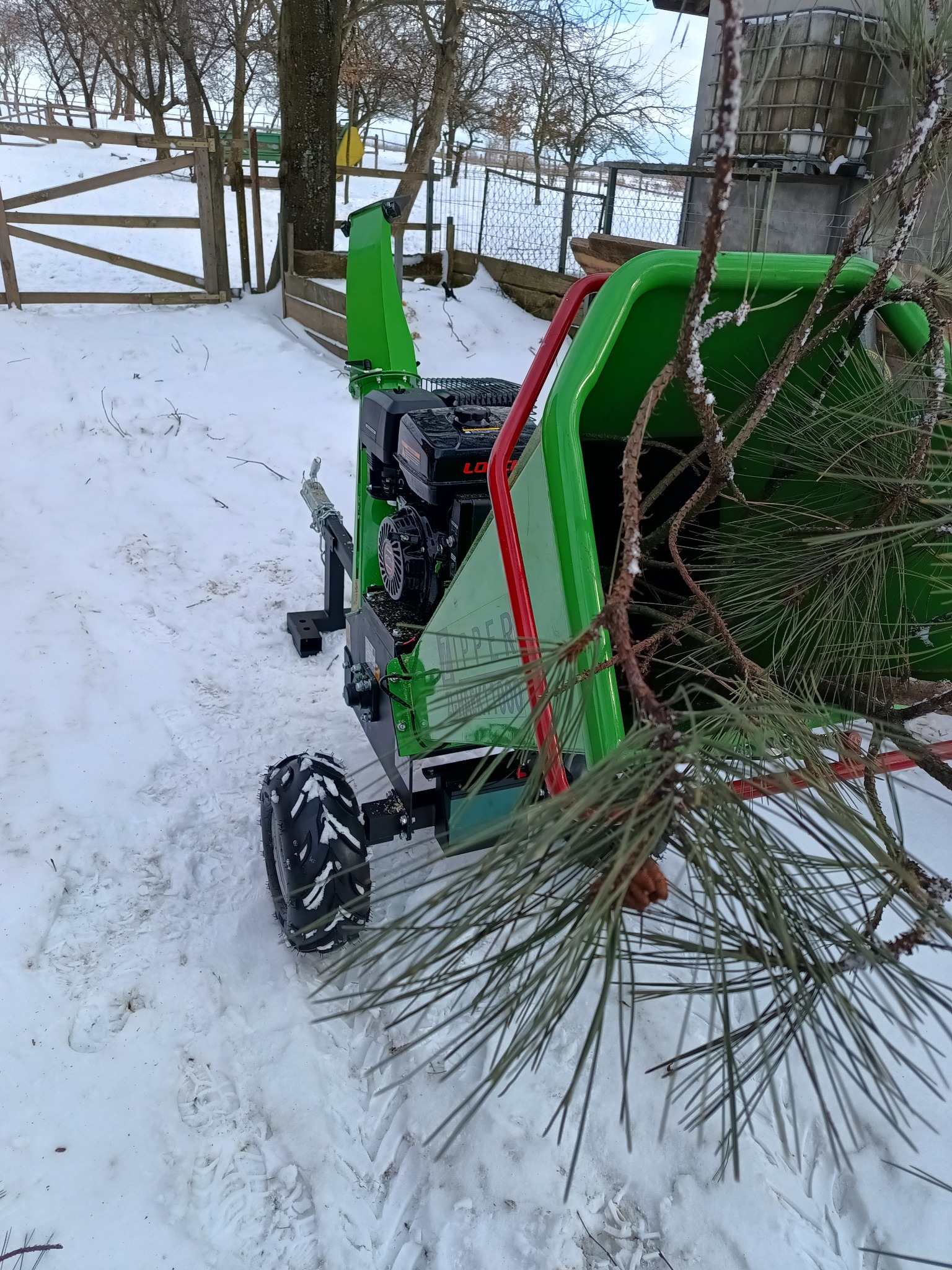 Zielona rębarka do gałęzi w zimowym krajobrazie, z gałęziami drzew iglastych w podajniku, śnieg na ziemi i drzewach w tle.