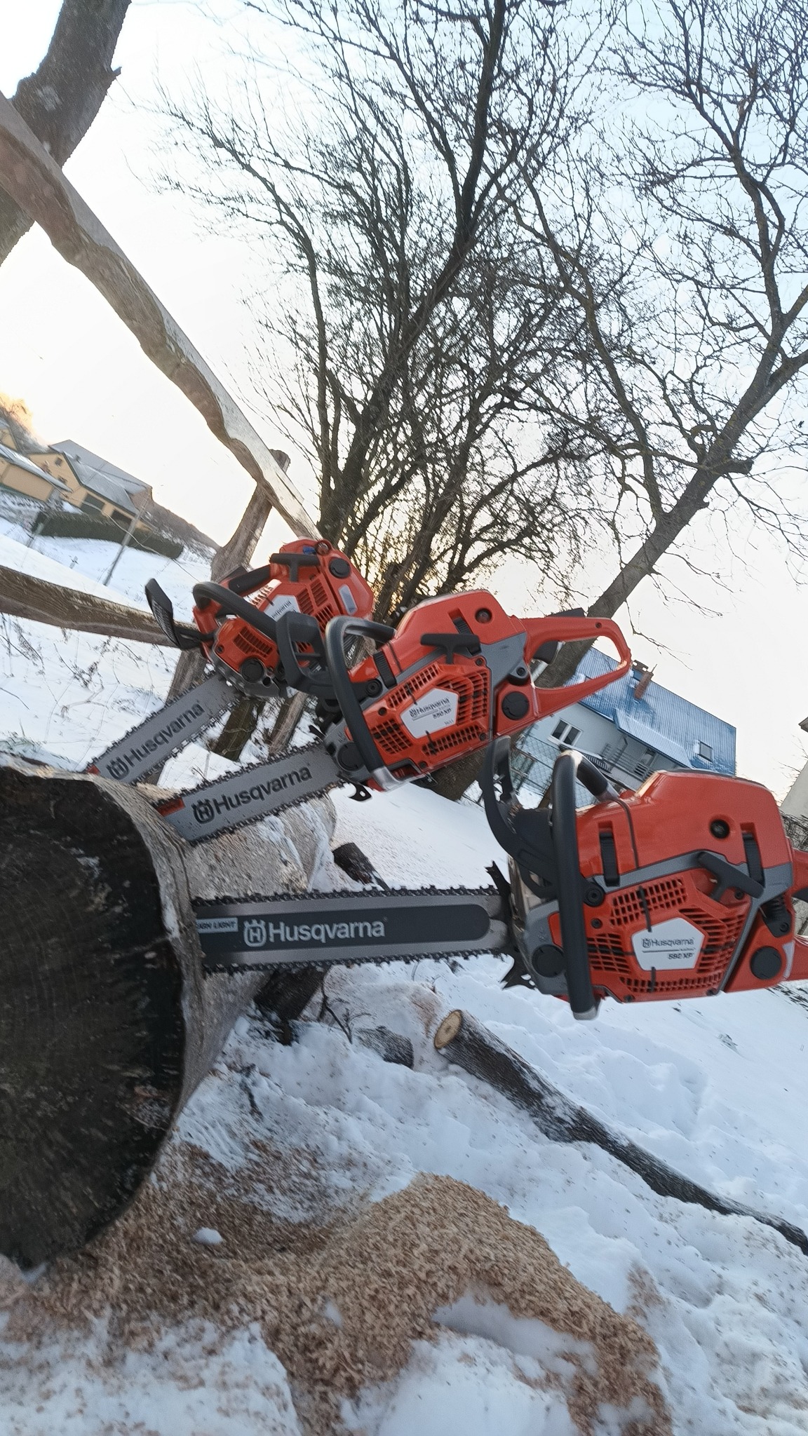 Trzy pomarańczowe pilarki Husqvarna spoczywają na kłodzie i śniegu, w tle drzewa bez liści i budynki.